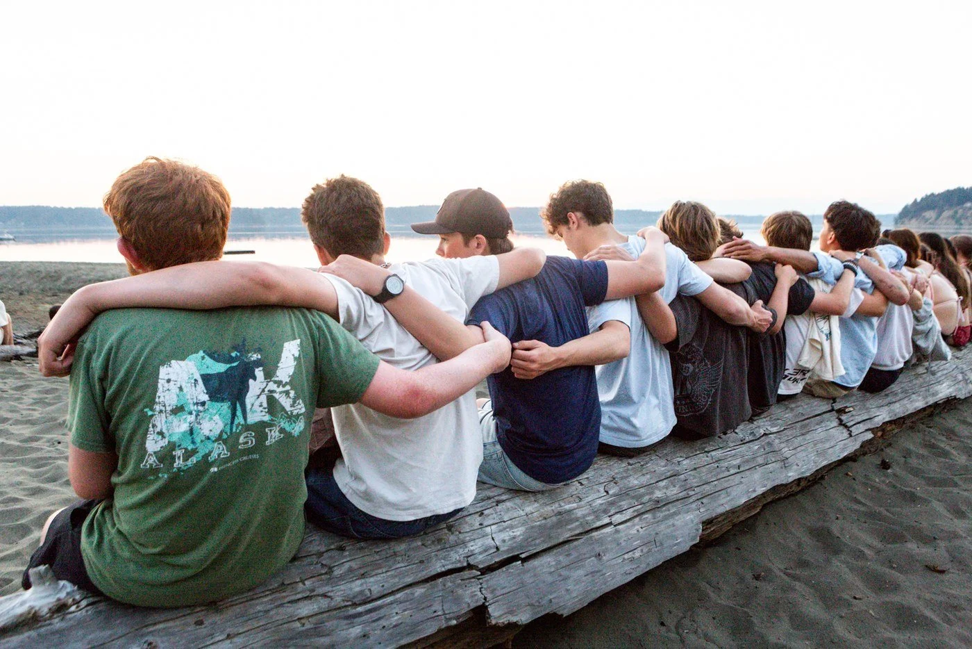A long row of campers sitting on a driftwood log on the beach at sunset, arms around each other, looking out at the water