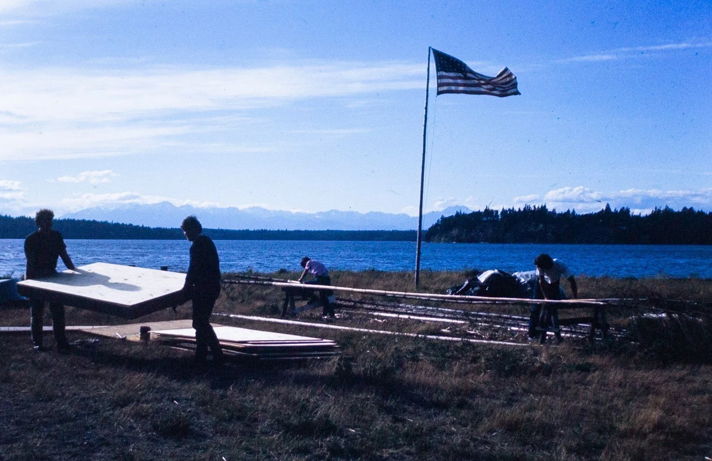 People carrying boards beside a flagpole on the shore in 1970, with the water and mountains behind