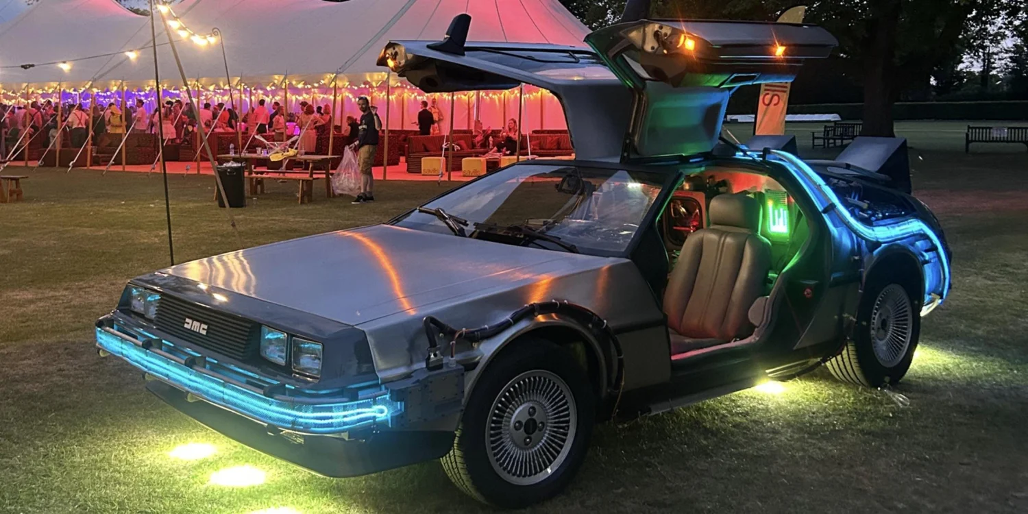 The time machine DeLorean with gull-wings up and blue underglow beside a lit marquee party at dusk