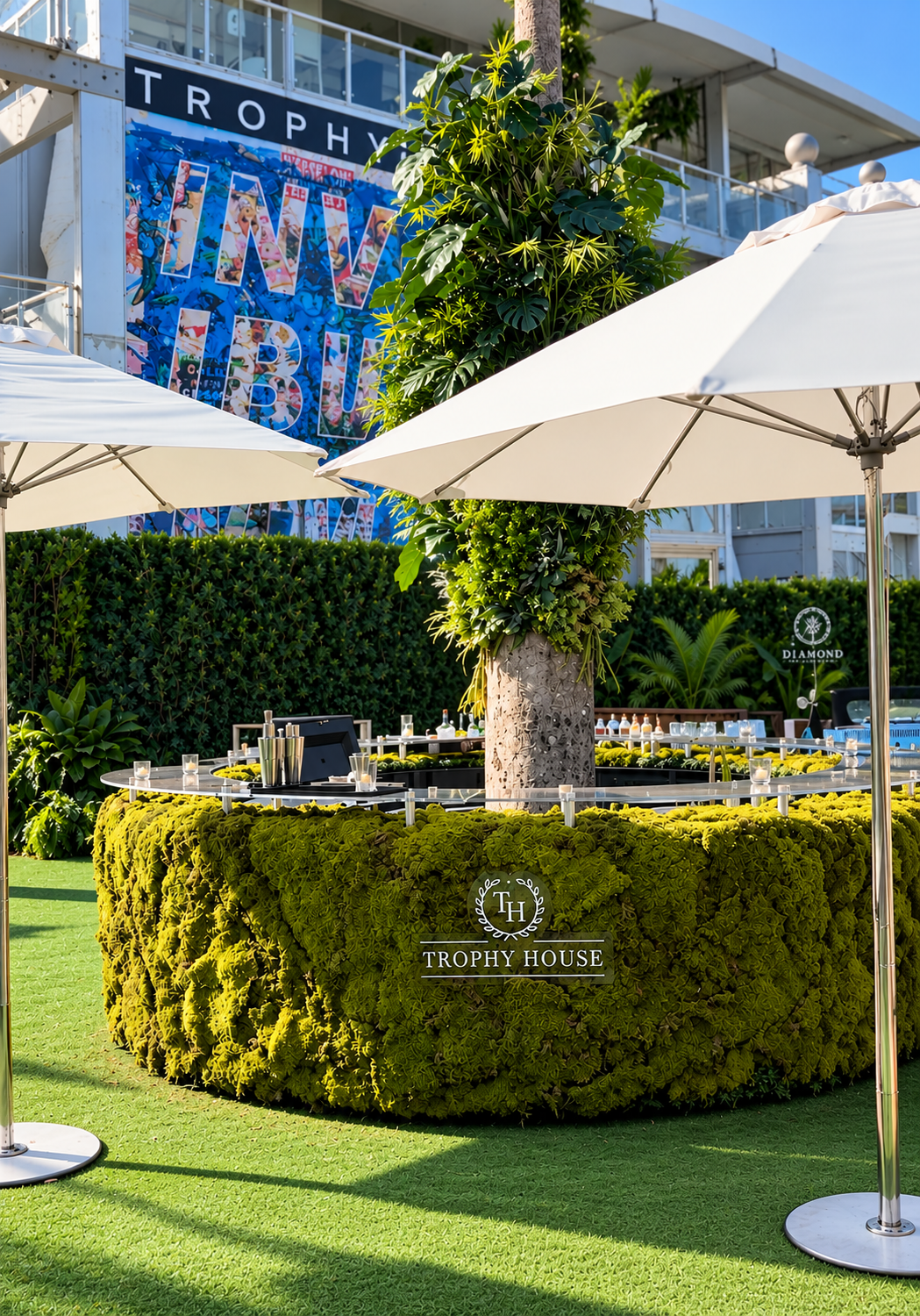 Moss-clad circular VIP bar with Trophy House logo wrapped around a foliage-covered palm trunk, under white umbrellas at Formula 1 Miami