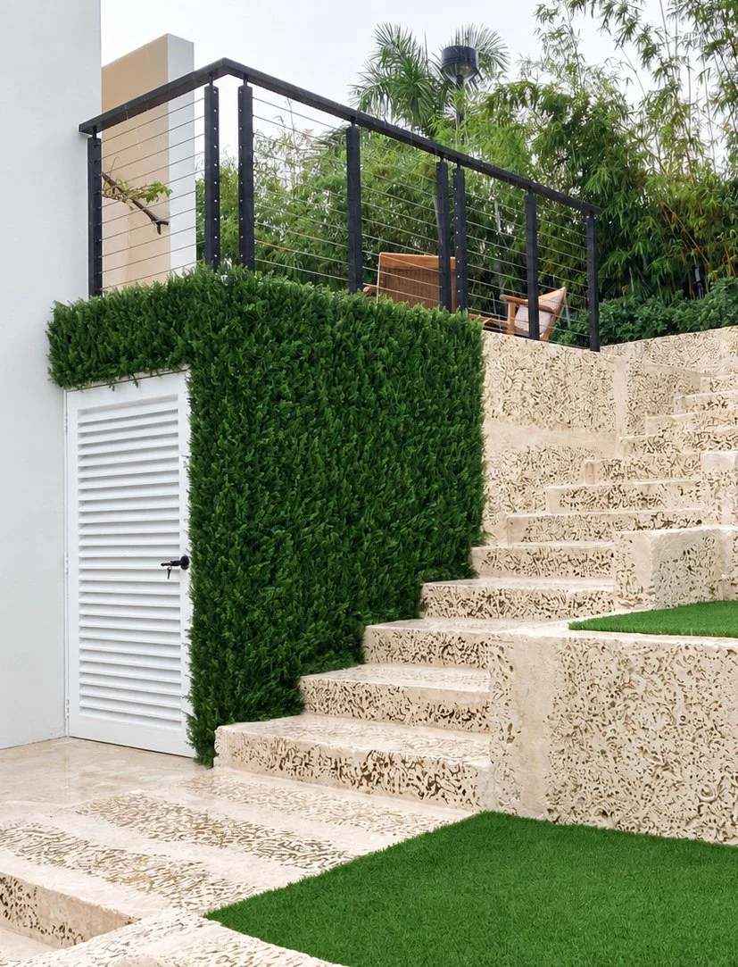Dense artificial hedge wall wrapping a white utility door beside coral stone stairs at a private residence in Coral Gables, FL