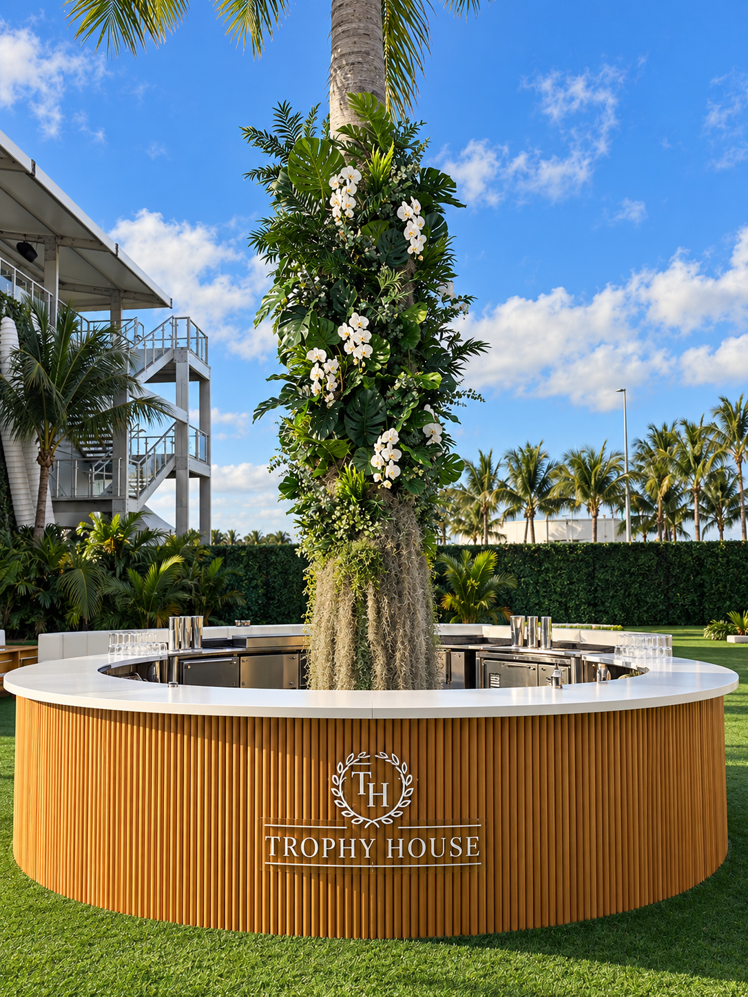 Palm trunk wrapped in layered tropical foliage and white orchids rising from the center of the round Trophy House bar at Formula 1 Miami