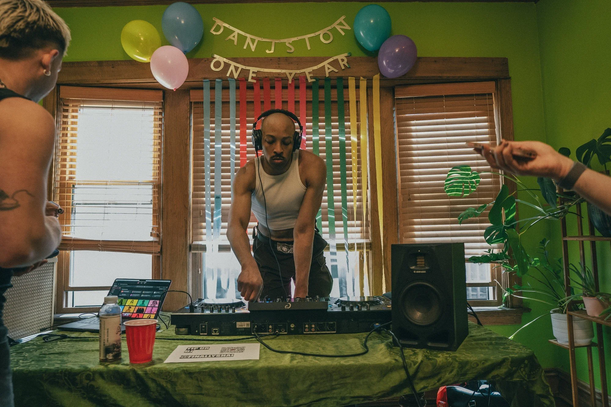 A DJ wearing headphones plays music beneath balloons and a party banner.