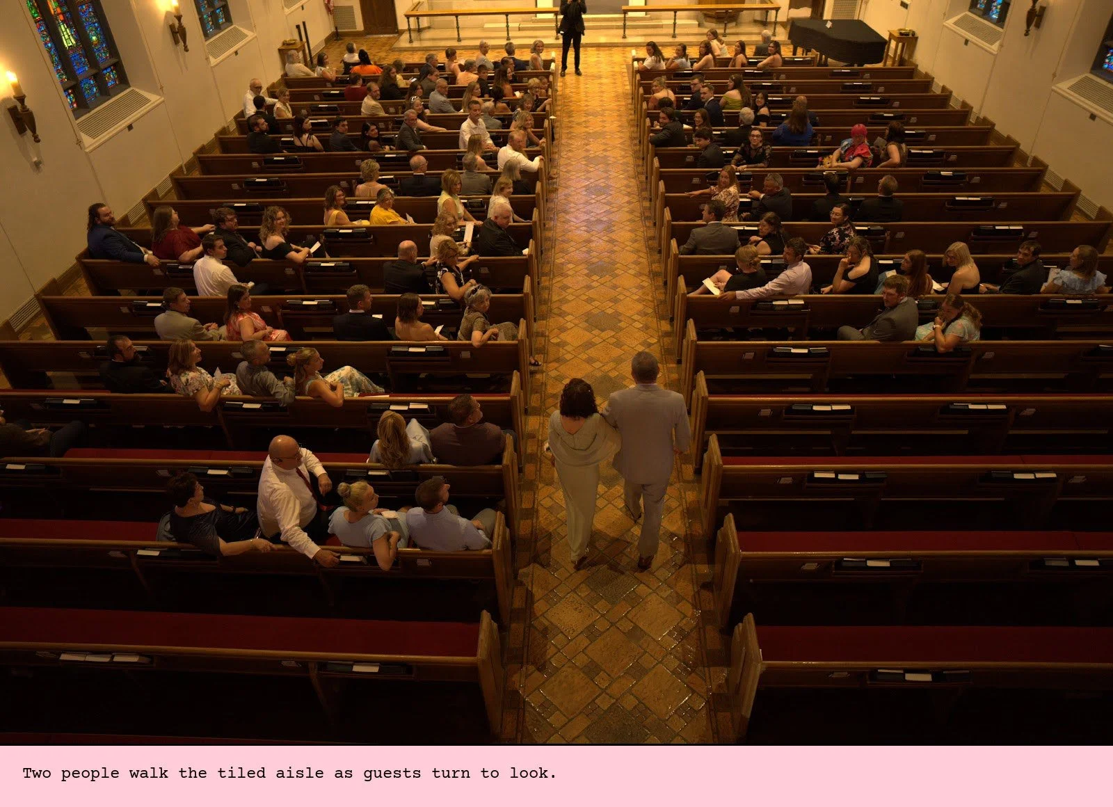 Two people walk the tiled aisle as guests turn to look.
