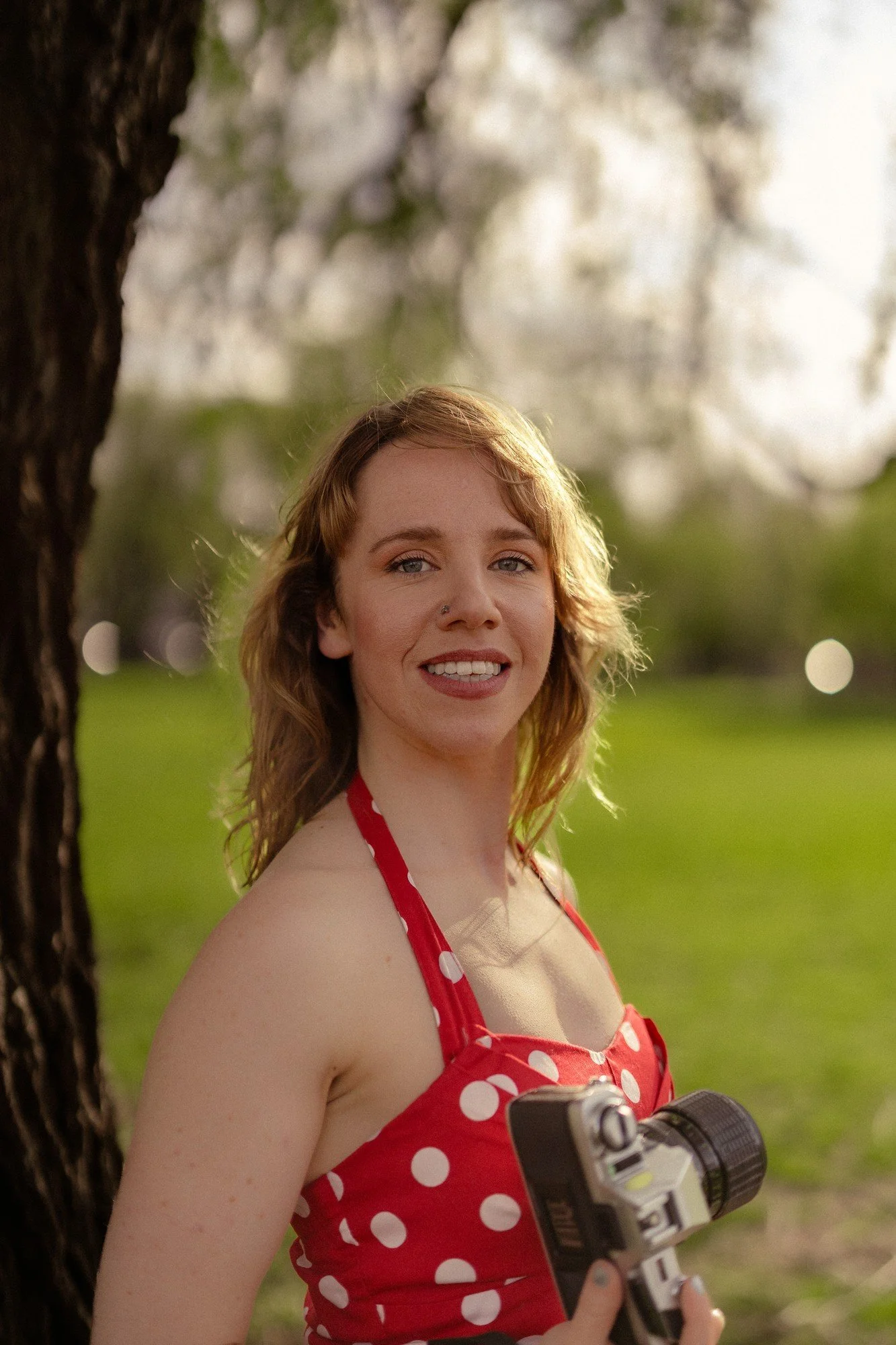 A person in a red polka-dot dress smiles beside a tree while holding a camera.