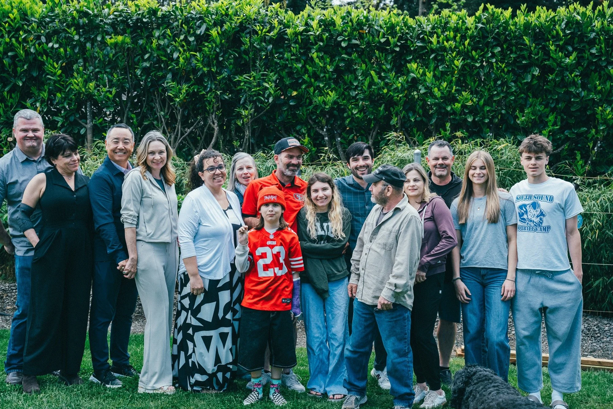 An extended family poses together in front of a leafy hedge.