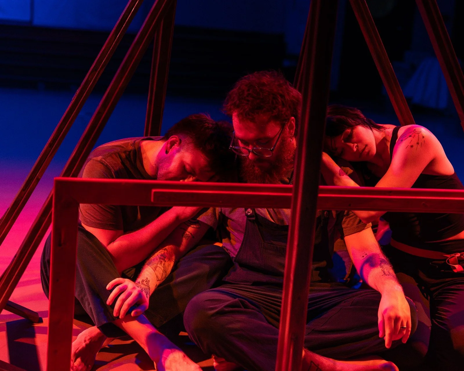 Three performers lean together inside a wooden house-shaped frame under red lighting.