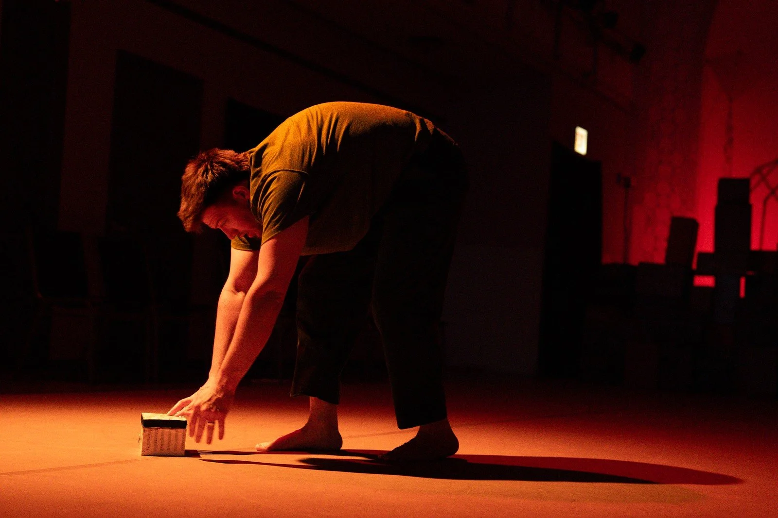 A performer bends toward a small object on the floor in a pool of warm light.