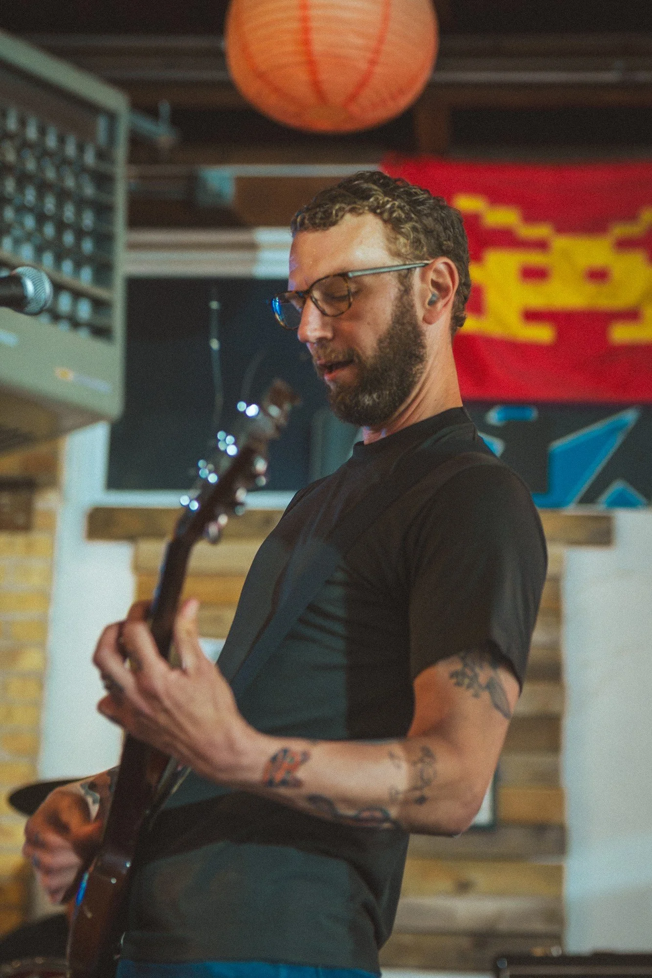 A guitarist in a black shirt plays beside a microphone, with a rainbow flag behind them.