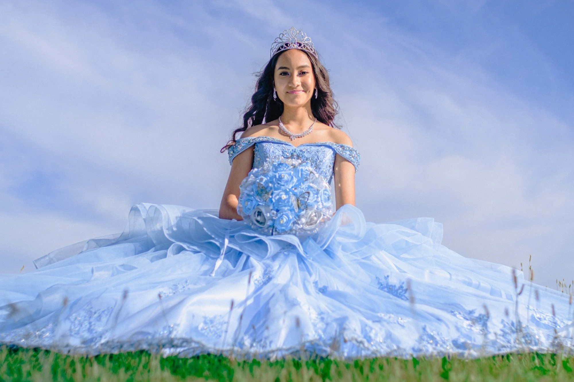 A person sits on the grass with a full blue gown spread around them beneath a cloudy sky.