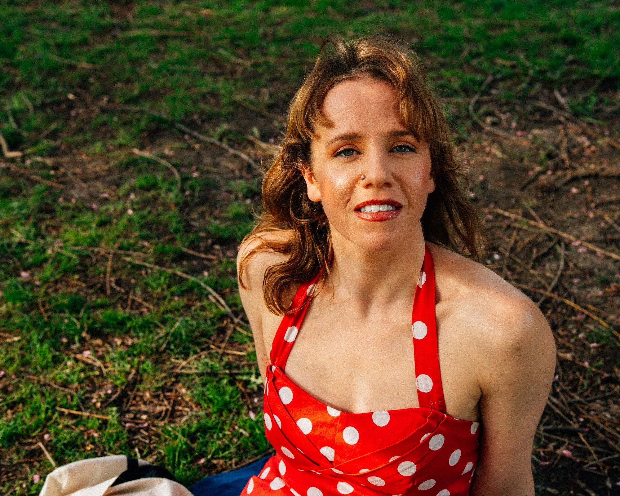 A person in a red polka-dot dress looks up toward the camera from a leafy patch of ground.