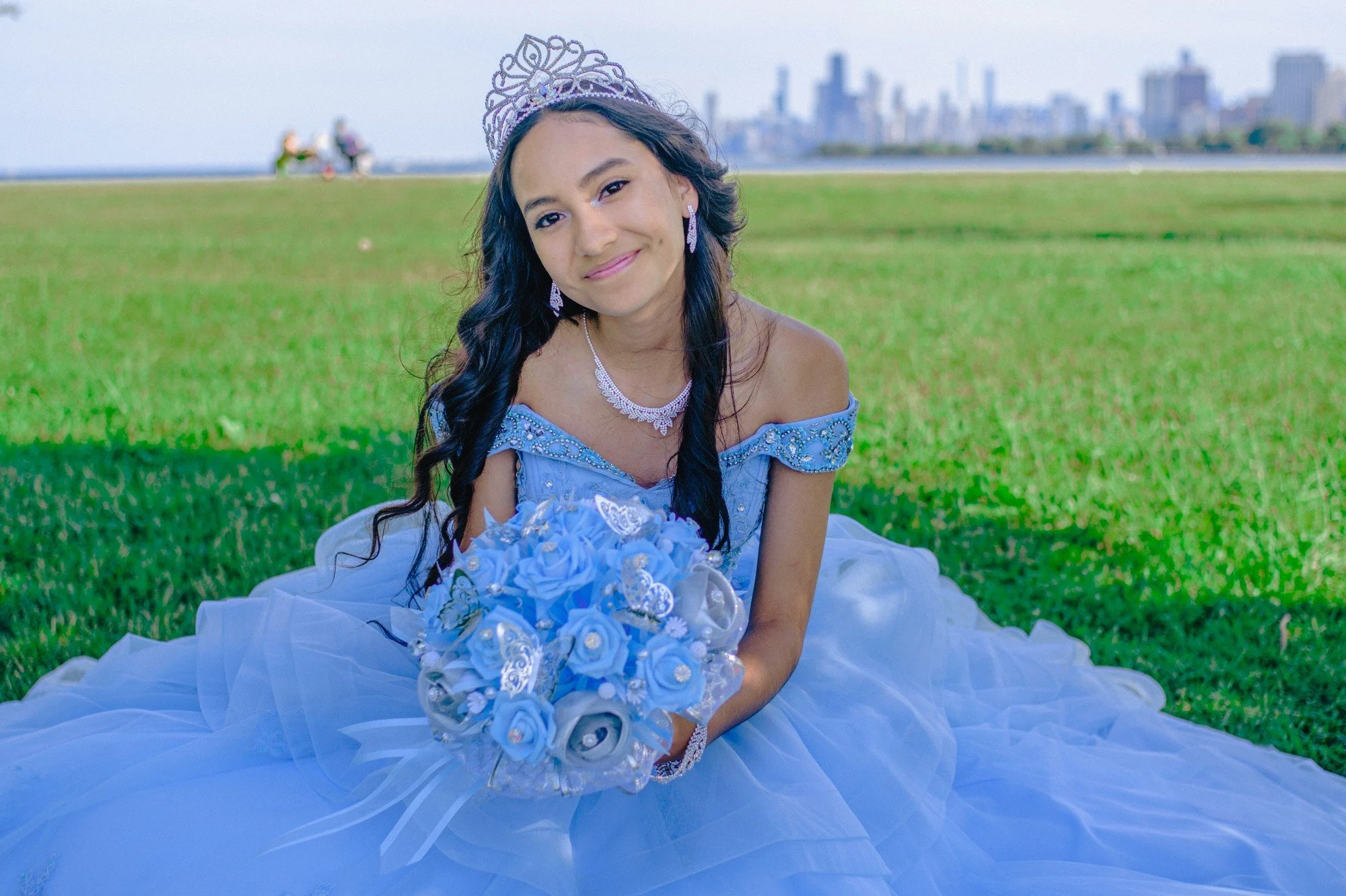 A person in a tiara leans toward the camera with a blue bouquet in their lap.