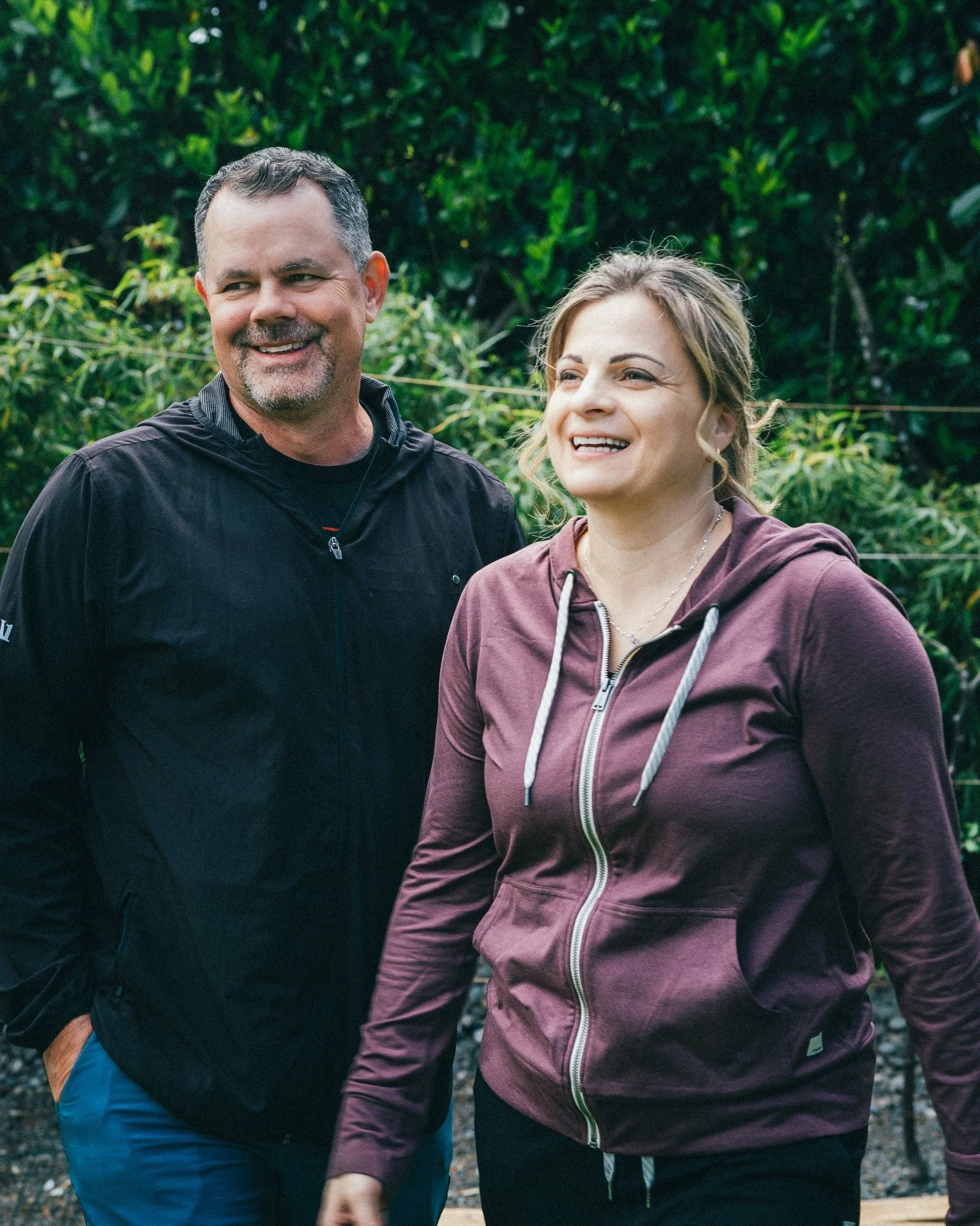 Two people laugh together beside a hedge.