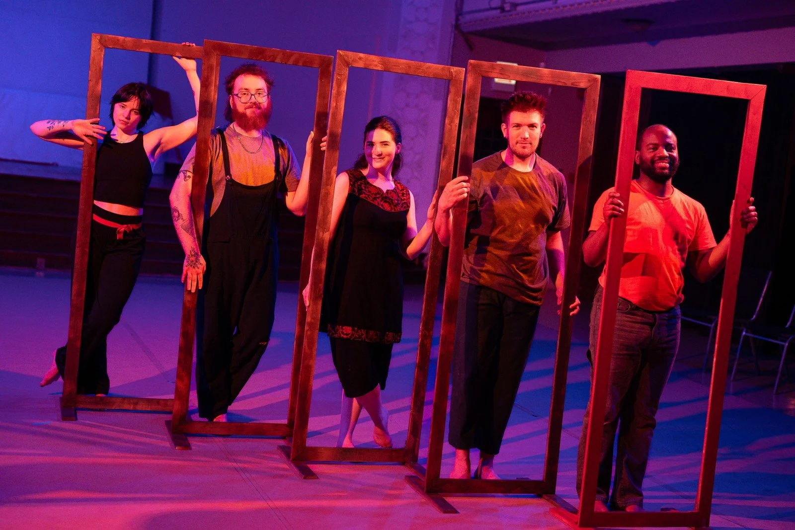 Five cast members pose inside wooden frames under purple stage lighting.