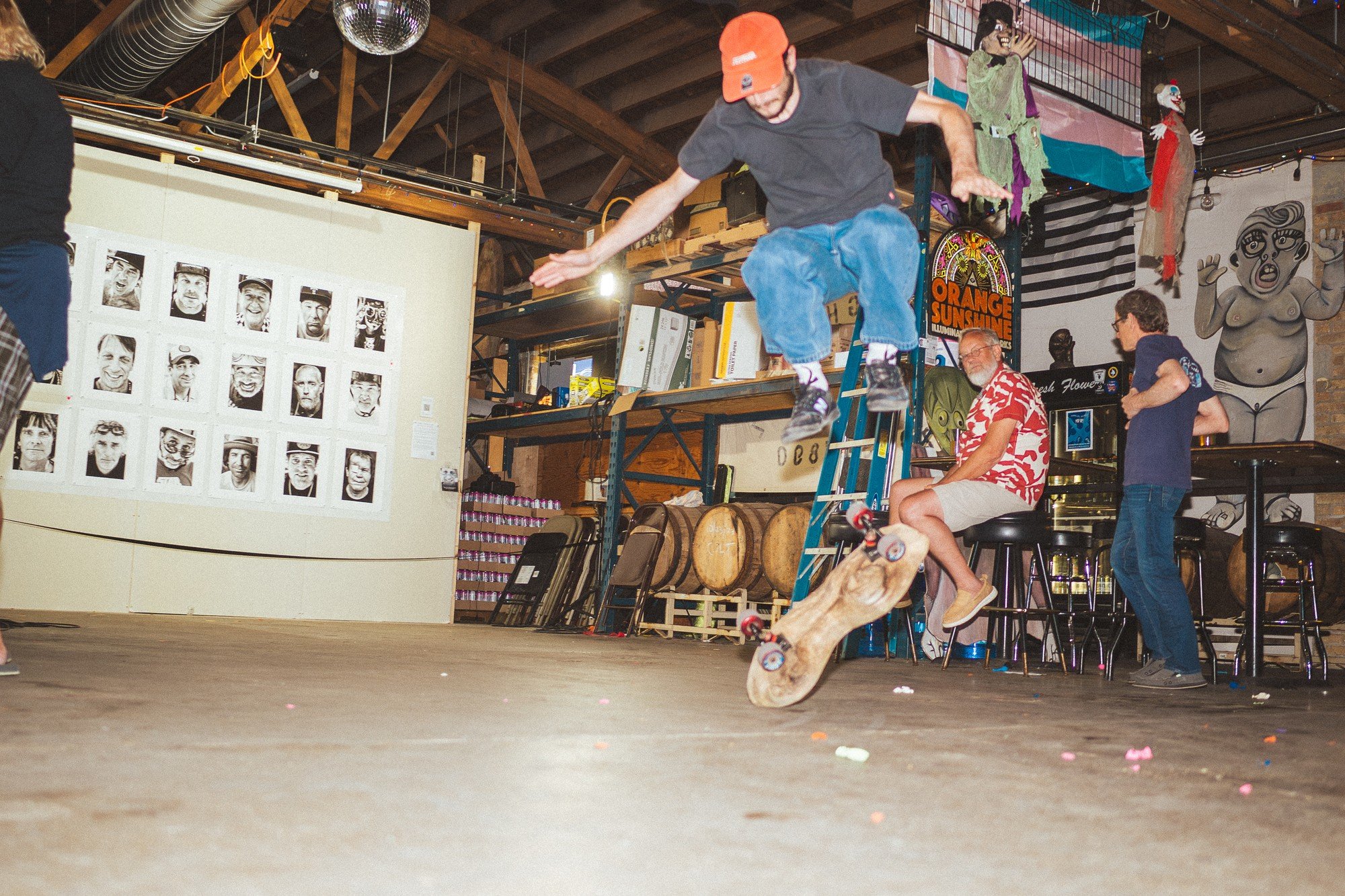 A skateboarder jumps with their board inside an art-filled room while onlookers watch.