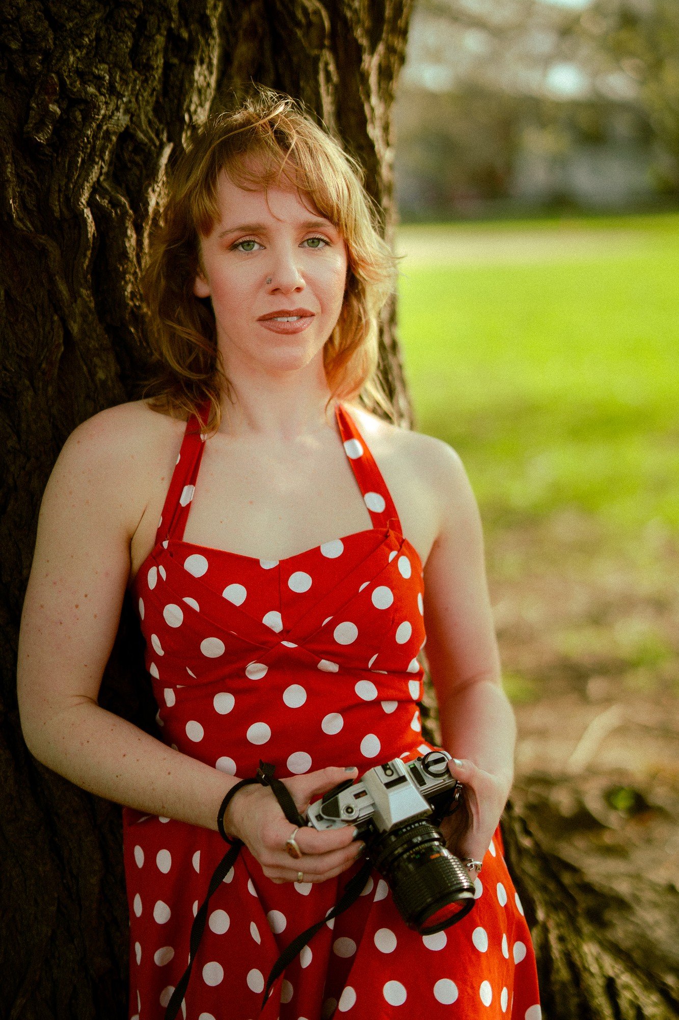 A person in a red polka-dot dress looks toward the camera beside a tree, holding their own camera at the waist.