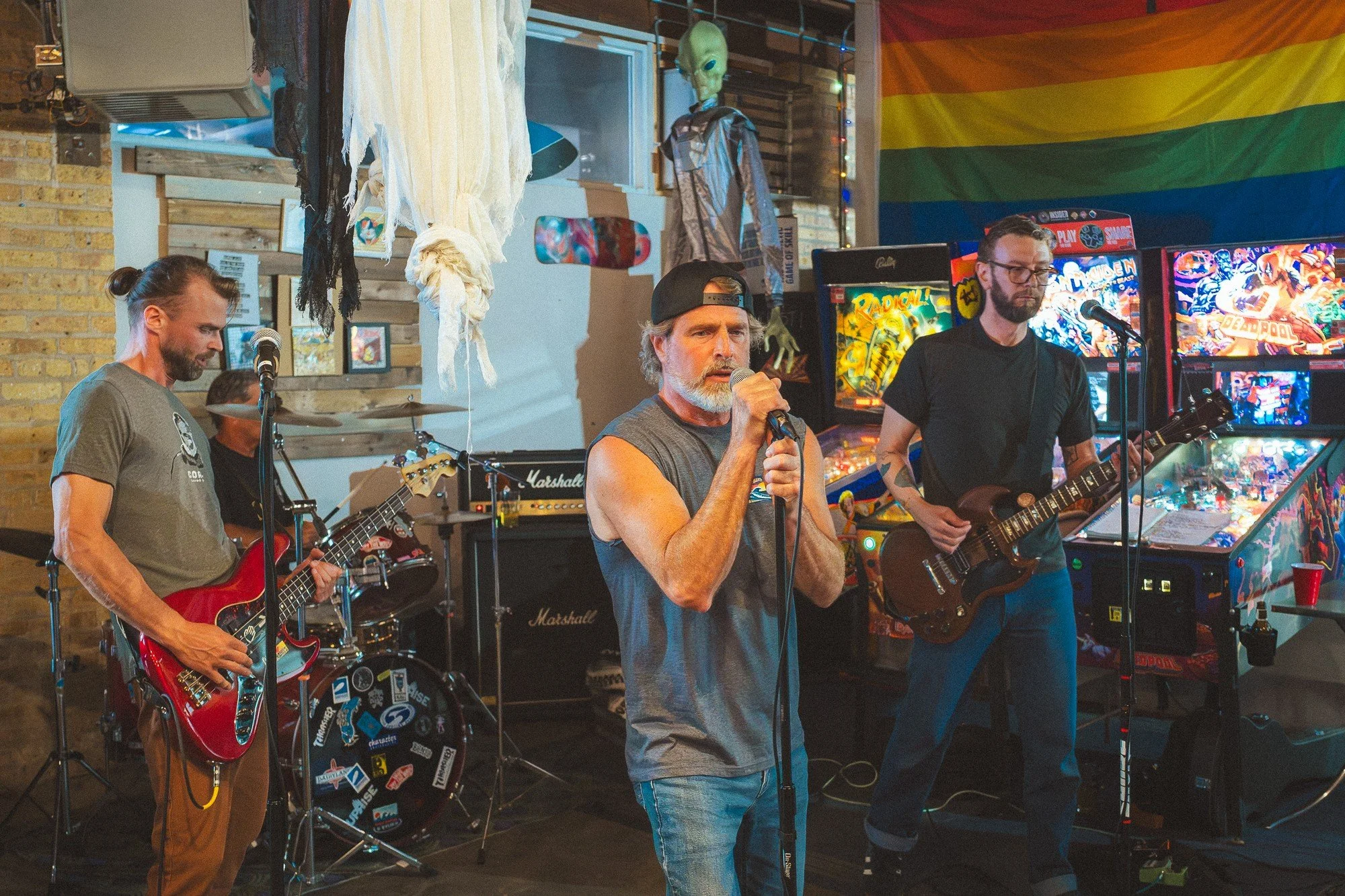 A vocalist sings between two guitarists, with pinball machines and a rainbow flag behind them.