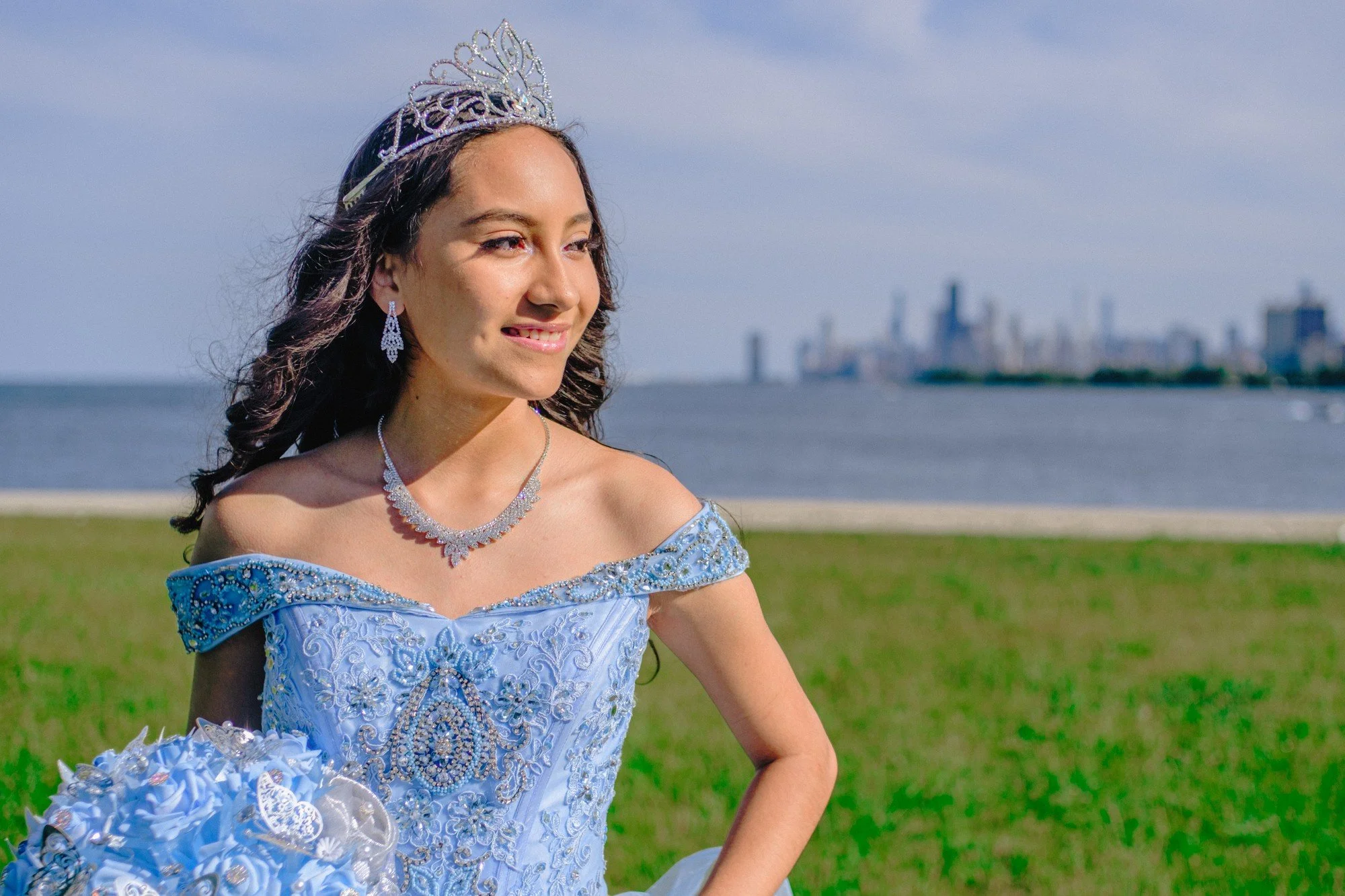 A smiling person in a blue gown and tiara holds a matching bouquet beside the lake.