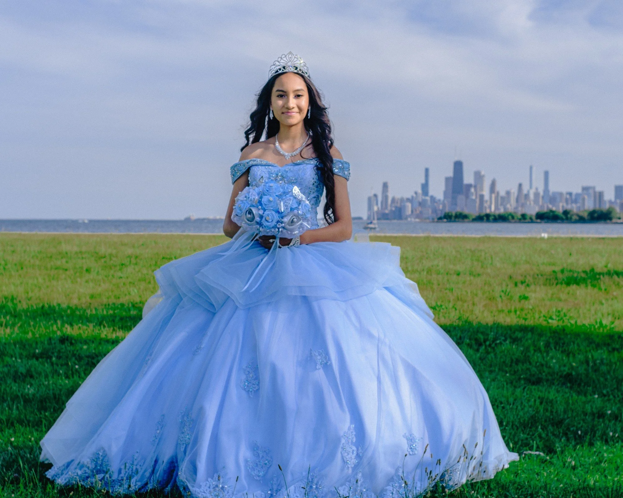 A person in a pale blue quinceañera gown stands on grass beside a lake, with the Chicago skyline behind them.