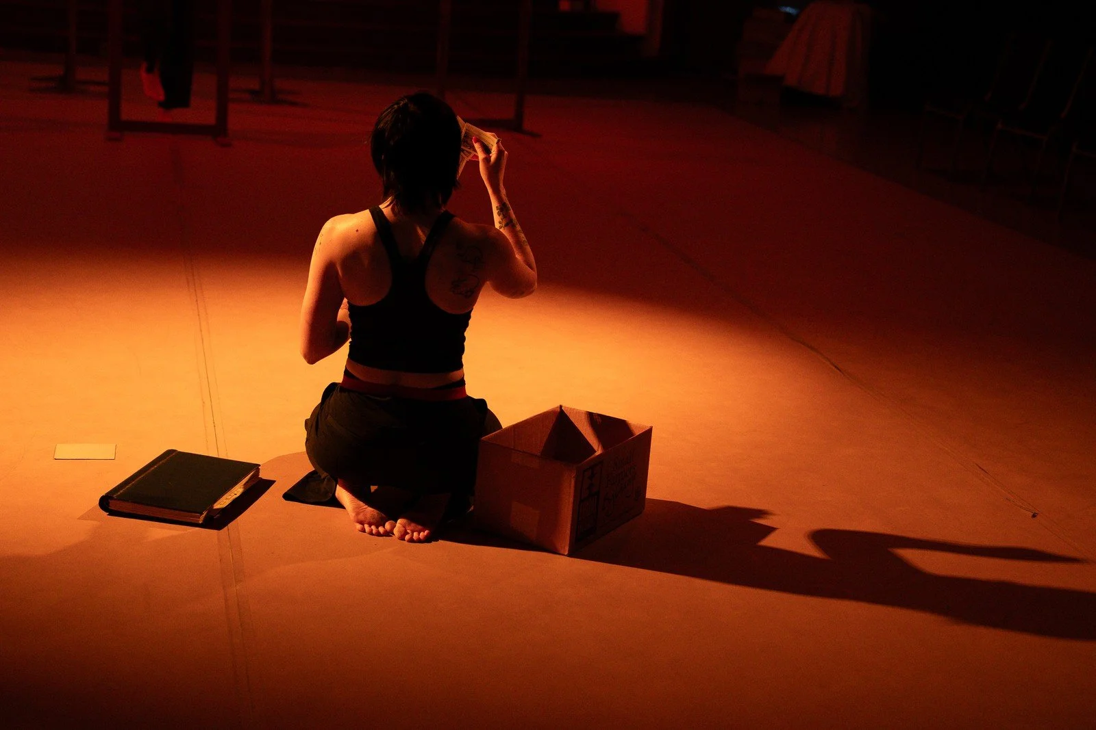 A seated performer and an open box cast a long shadow across an amber-lit floor.