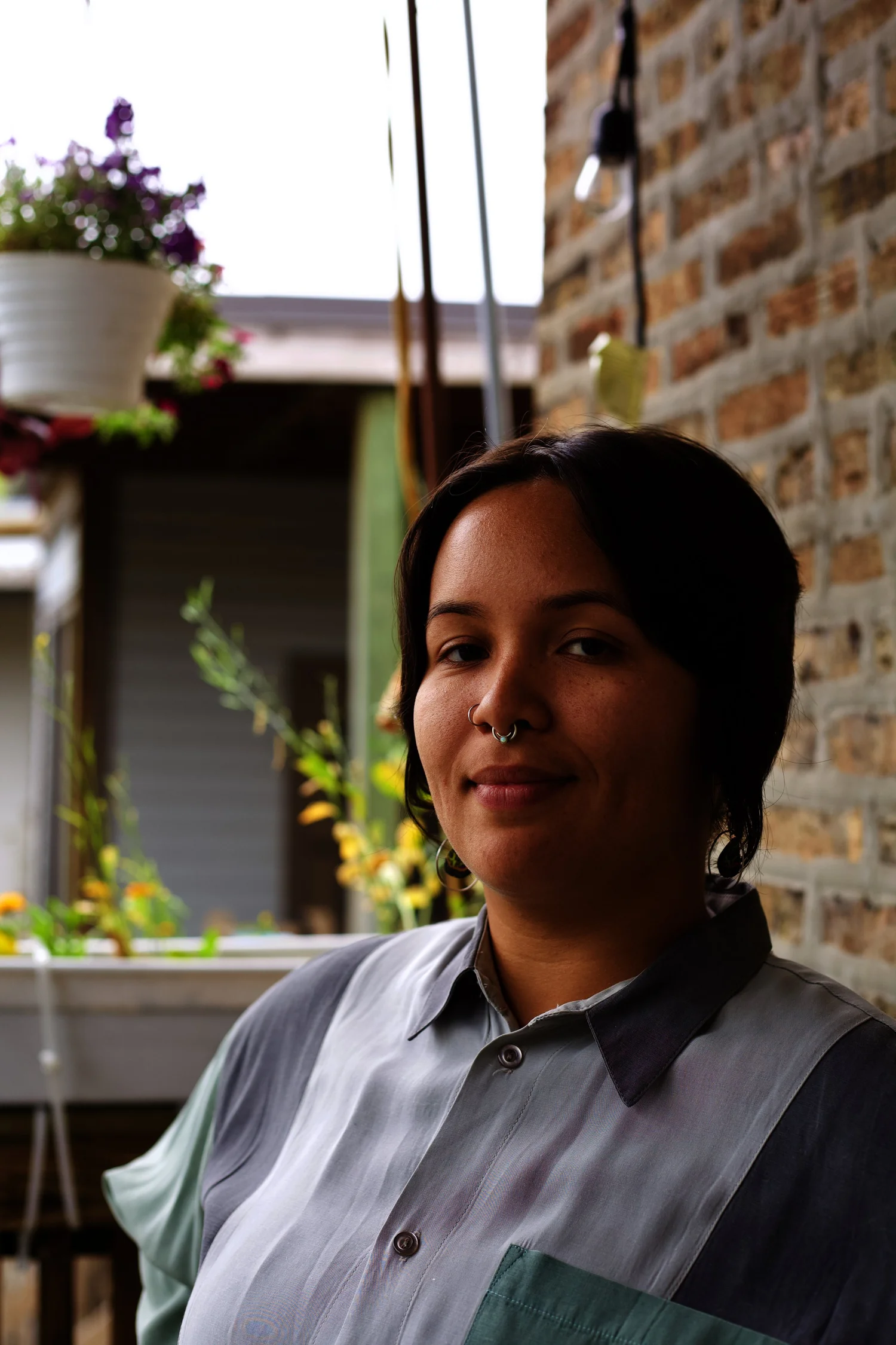A person in a gray-and-green shirt looks toward the camera beside a brick wall outdoors.