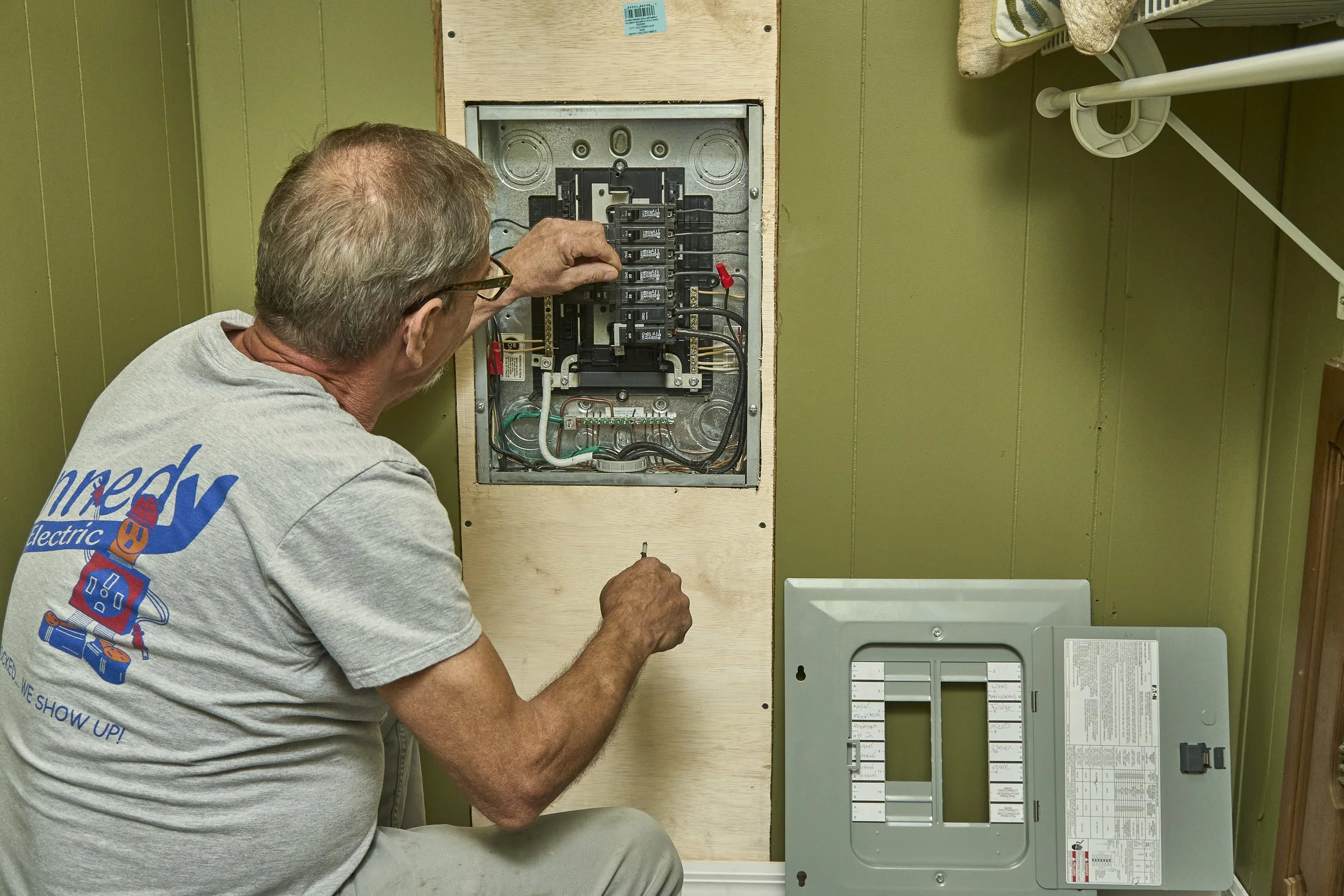 Electrician (Kennedy shirt) at an open residential breaker panel with the cover removed and set beside it