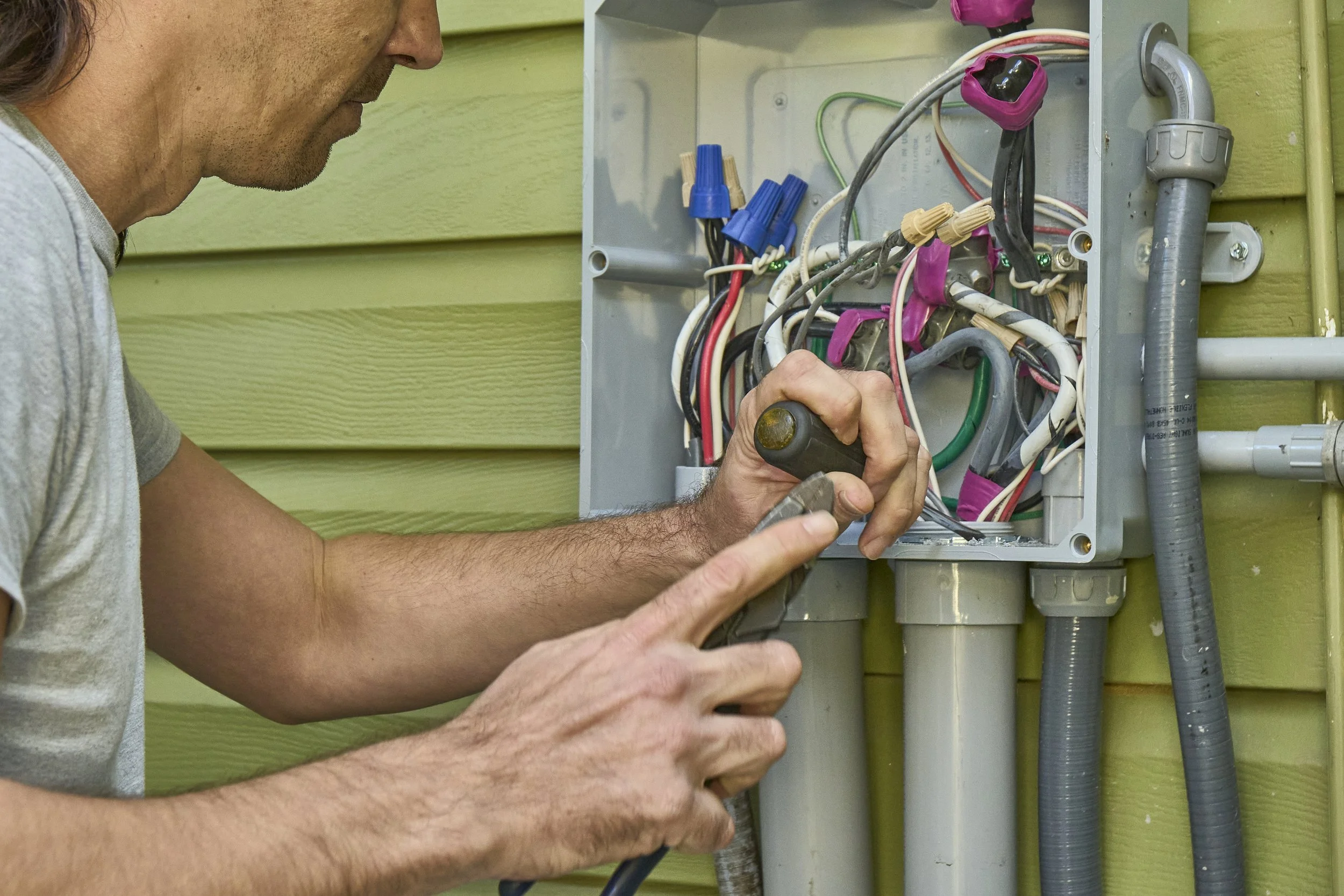 Hands working on an open exterior junction box with colorful wires on green siding