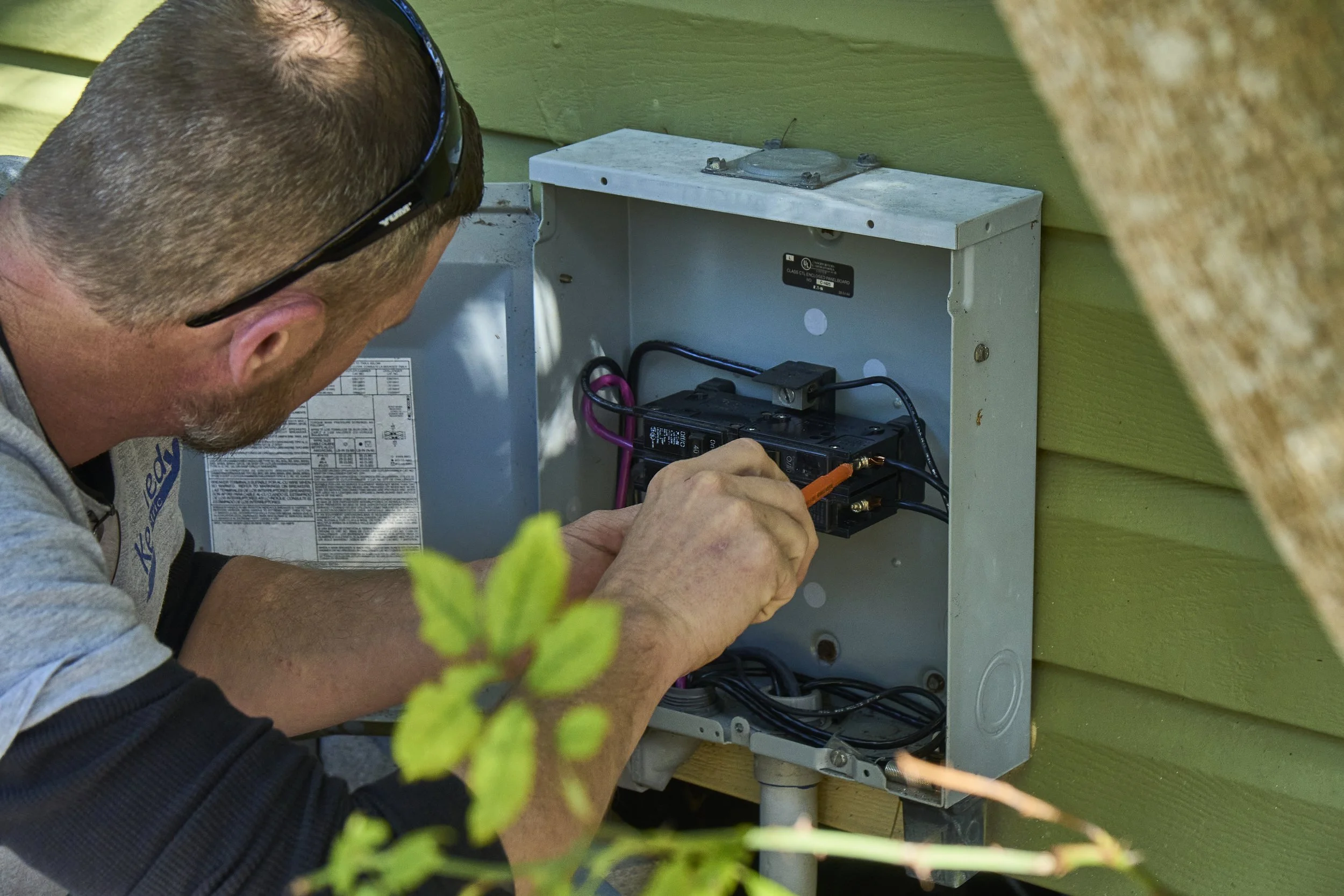 Man at an open exterior breaker/disconnect panel on green siding