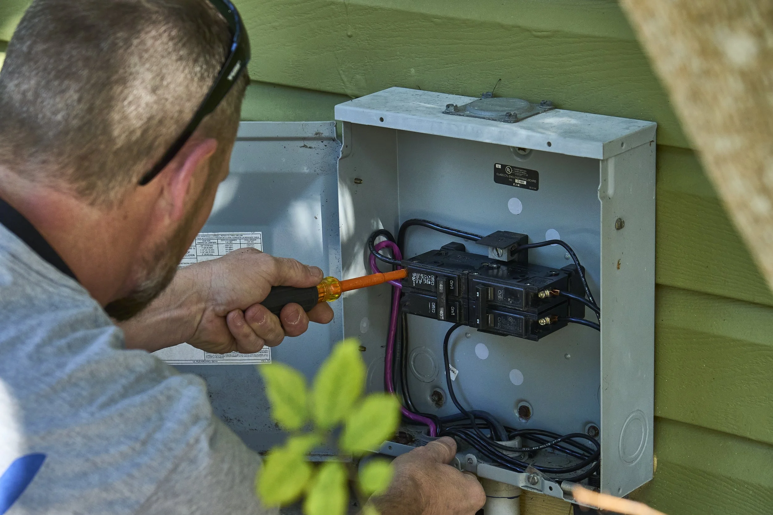 Man with a screwdriver at an open exterior breaker/disconnect panel on green siding