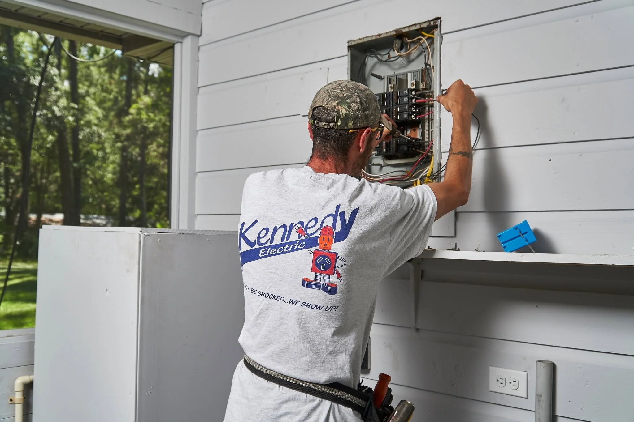 Electrician (back) working at an open exterior panel beside a fridge on a porch