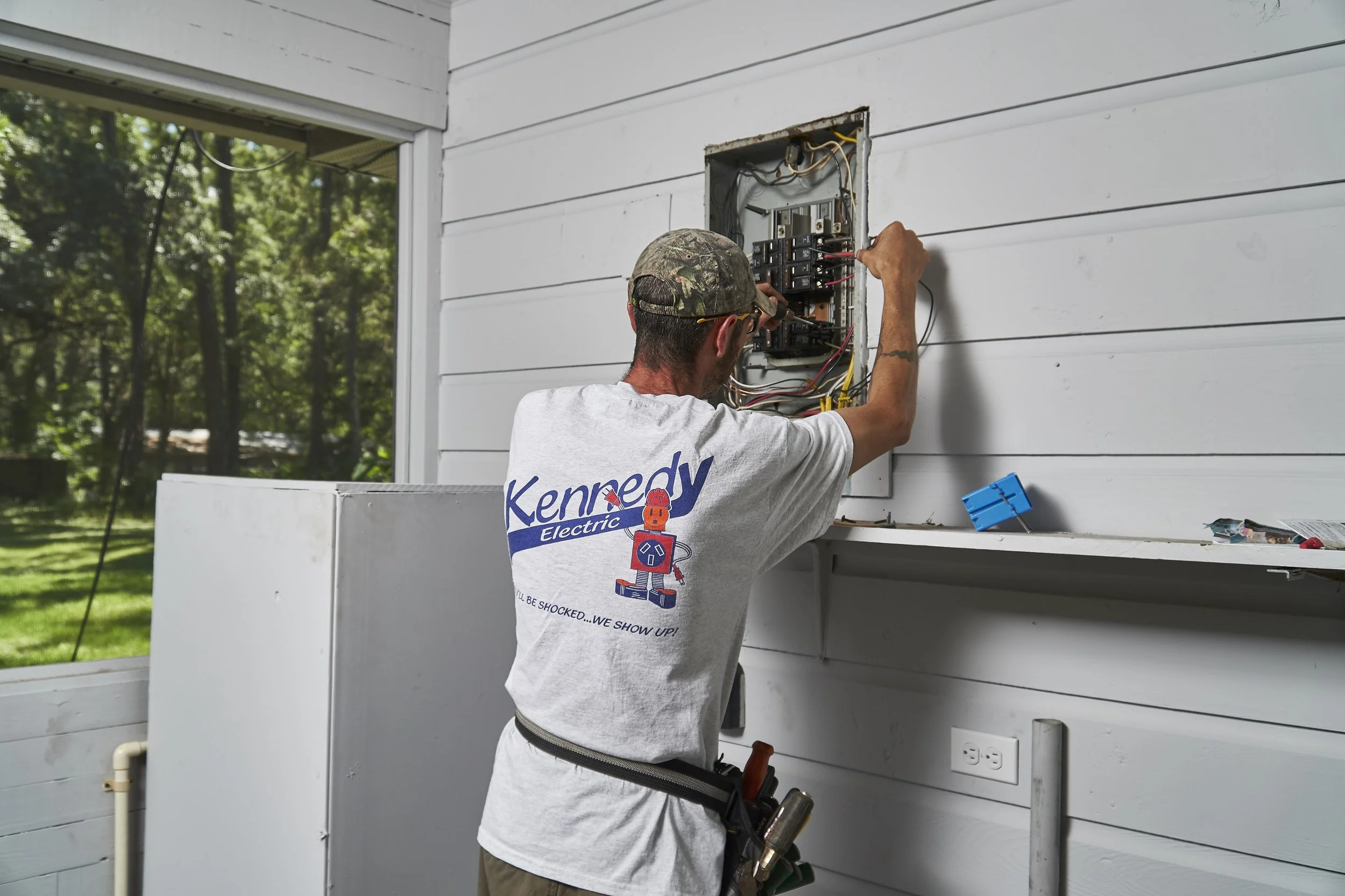 Electrician (back) working at an open interior panel with exposed wiring/breakers, tools on belt