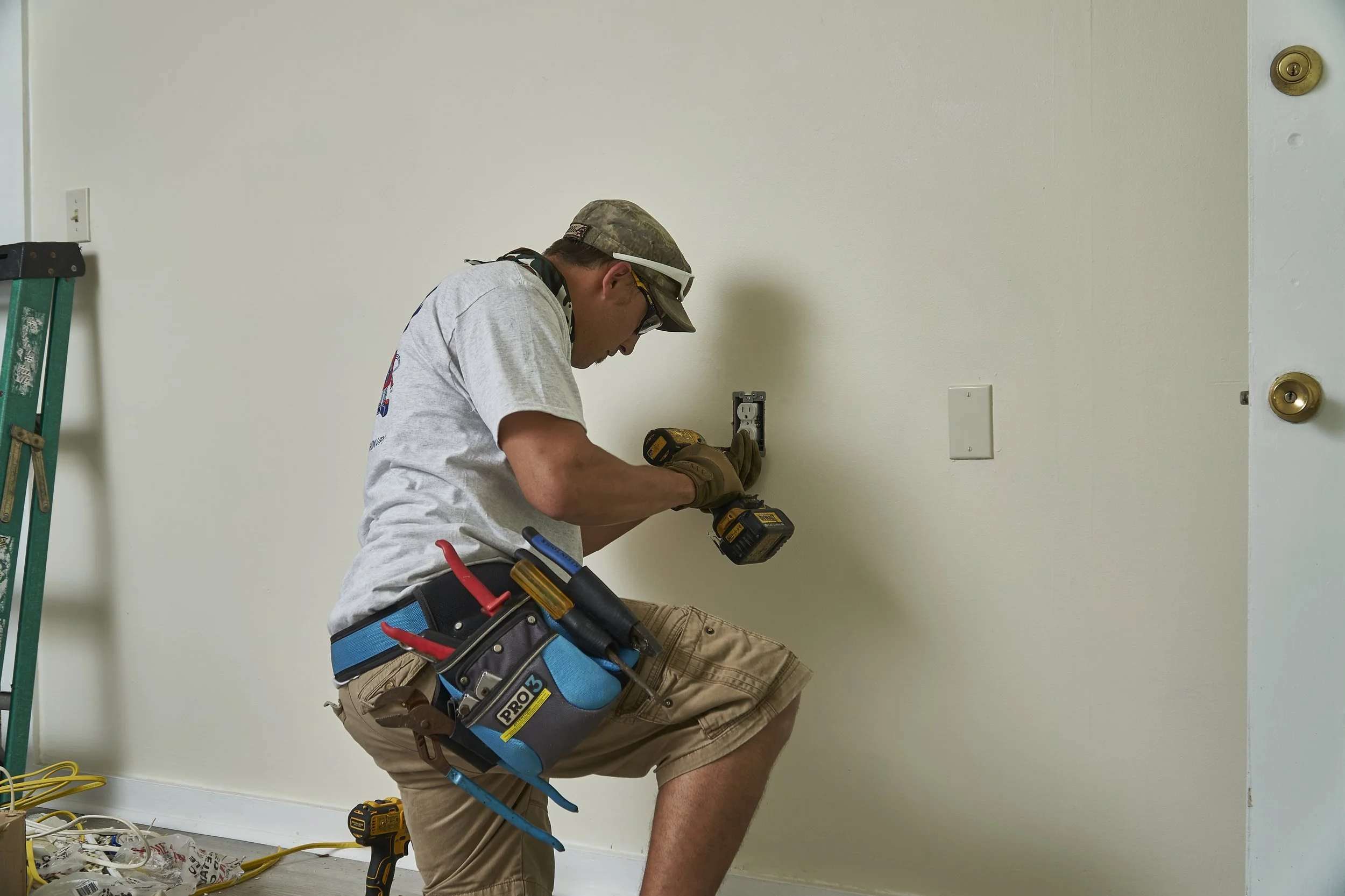 Electrician kneeling at an interior wall box, ladder and toolbag