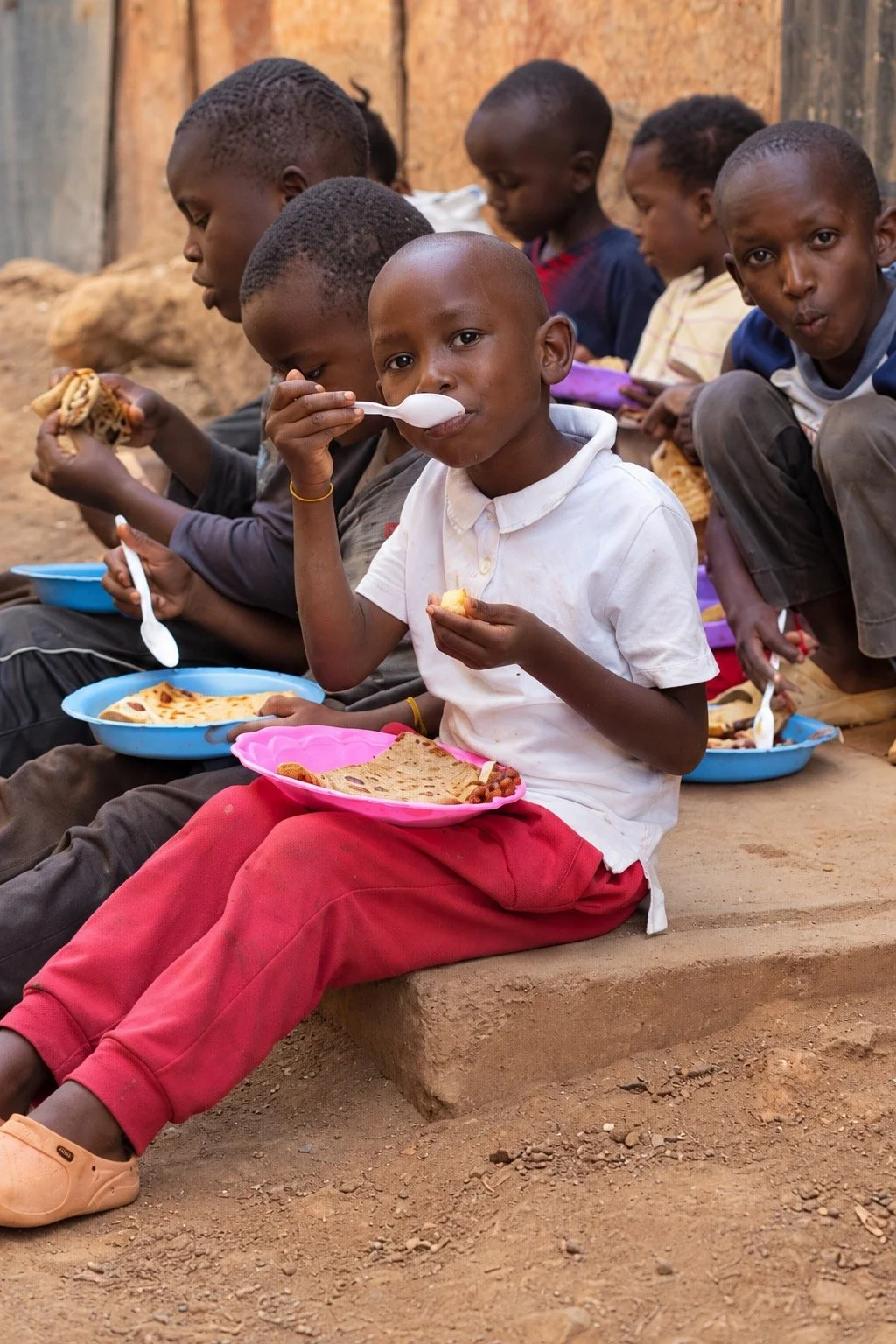 A little boy in a white shirt eats from a pink plate with other children in Nairobi.