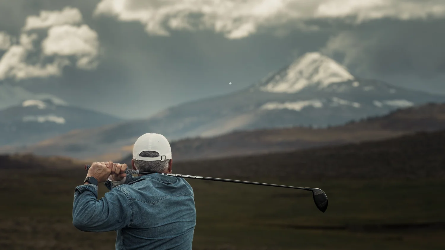 Closer rear view of a golfer holding the finish of a driver swing as the ball flies toward the mountains.
