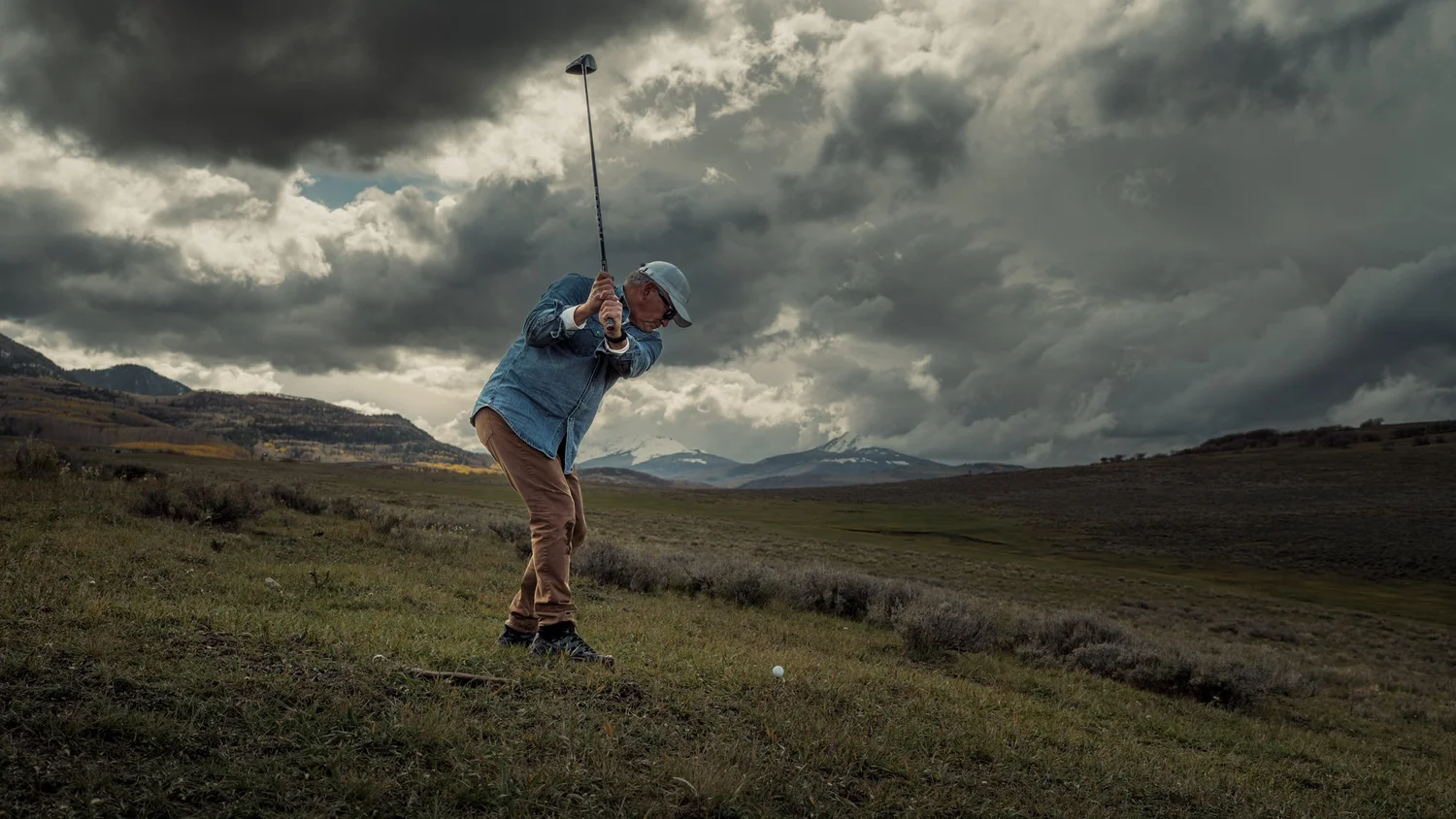 A golfer at the top of a driver backswing on open range beneath towering clouds.