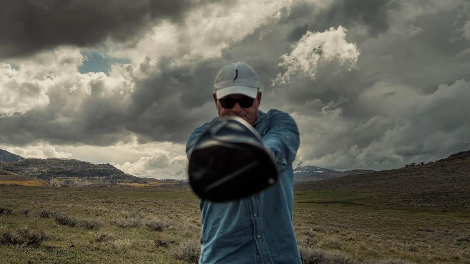 A golfer playfully points a driver toward the camera beneath dramatic clouds.