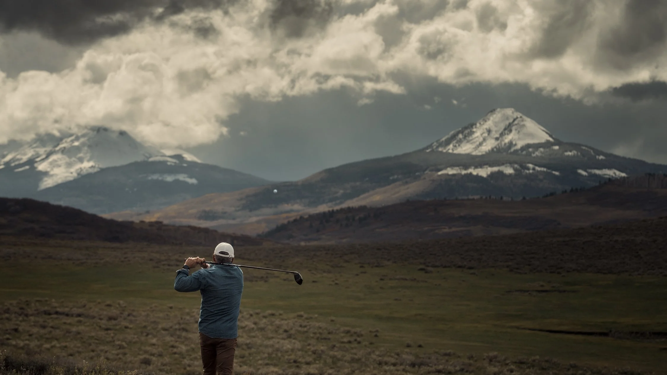 A golfer finishes a driver swing on open sagebrush range beneath snowy San Juan peaks, with the ball in flight.