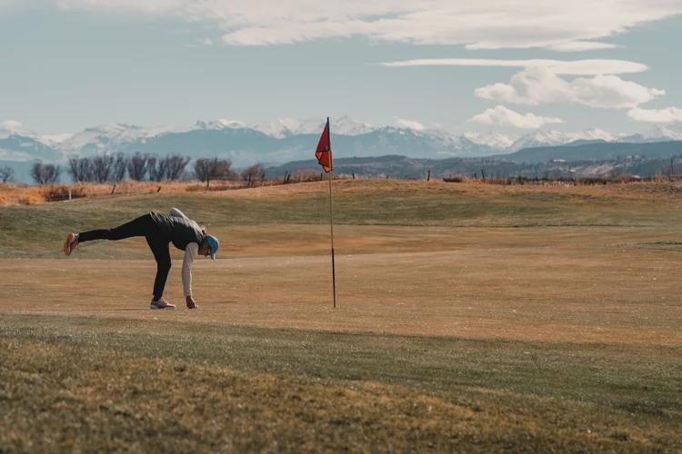 A golfer bends toward the green, with a red flag and mountains beyond.
