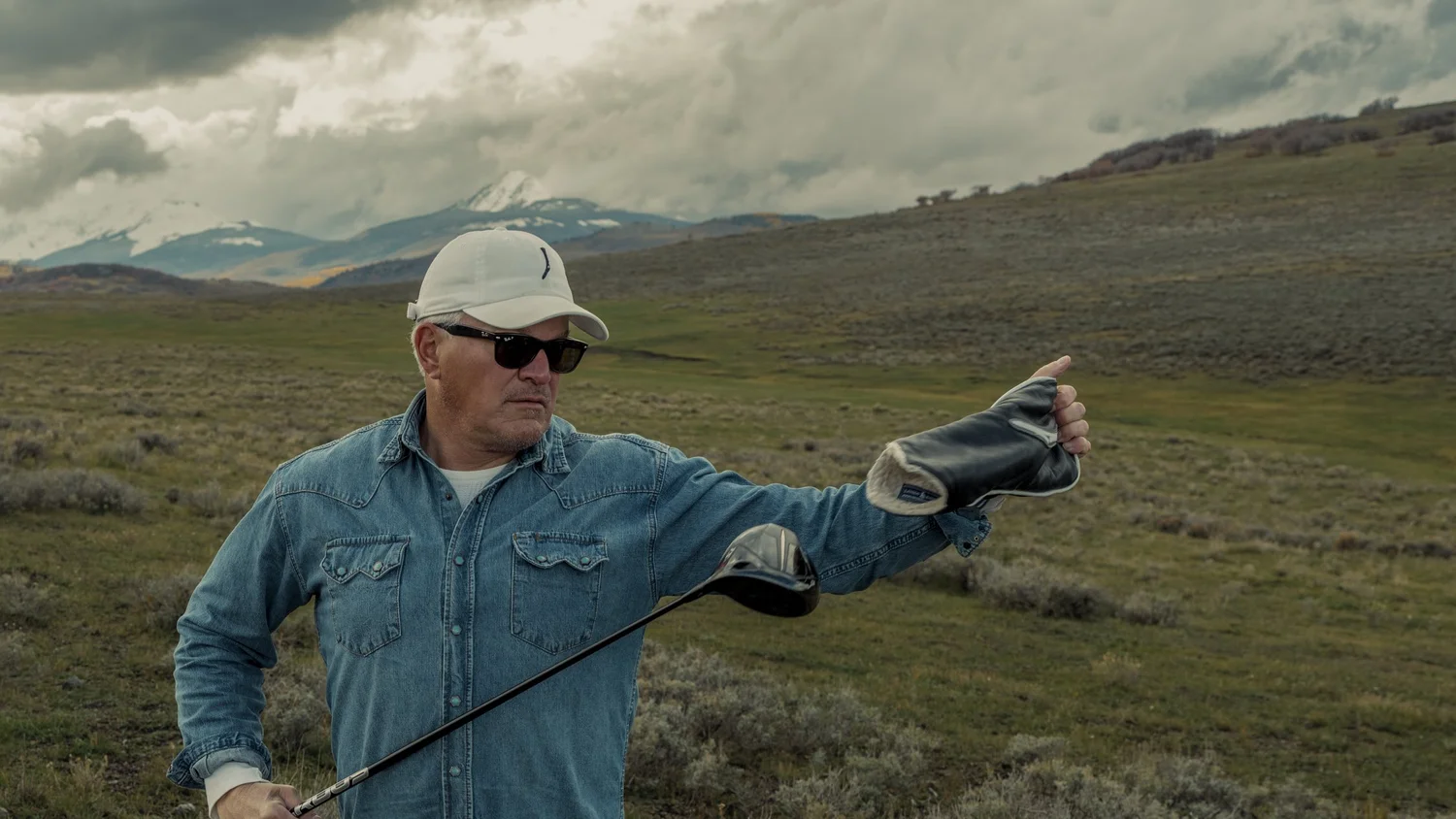 A golfer in a denim Western shirt removes the headcover from a driver in the Colorado landscape.