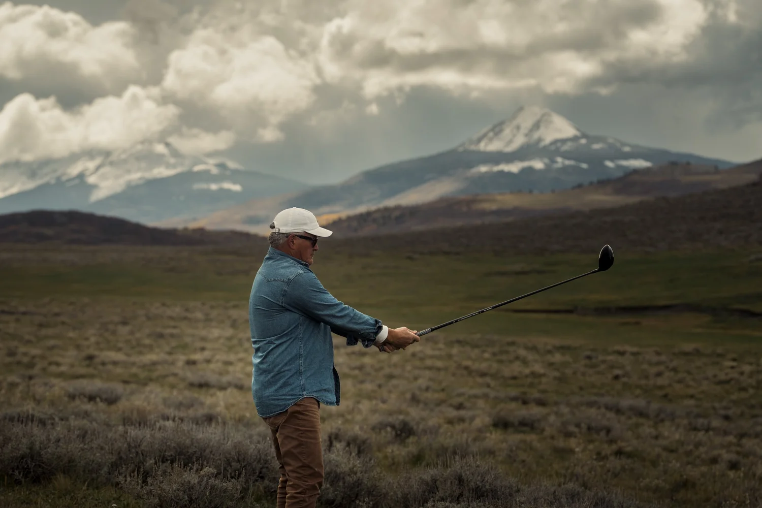 A golfer holds a driver forward with snowy San Juan peaks beyond.