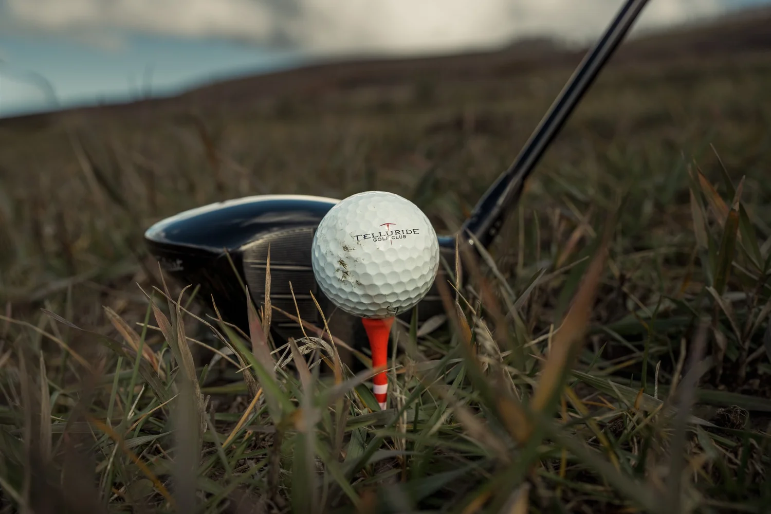 A Telluride Golf Club ball on an orange tee, with a driver resting behind it in native grass.