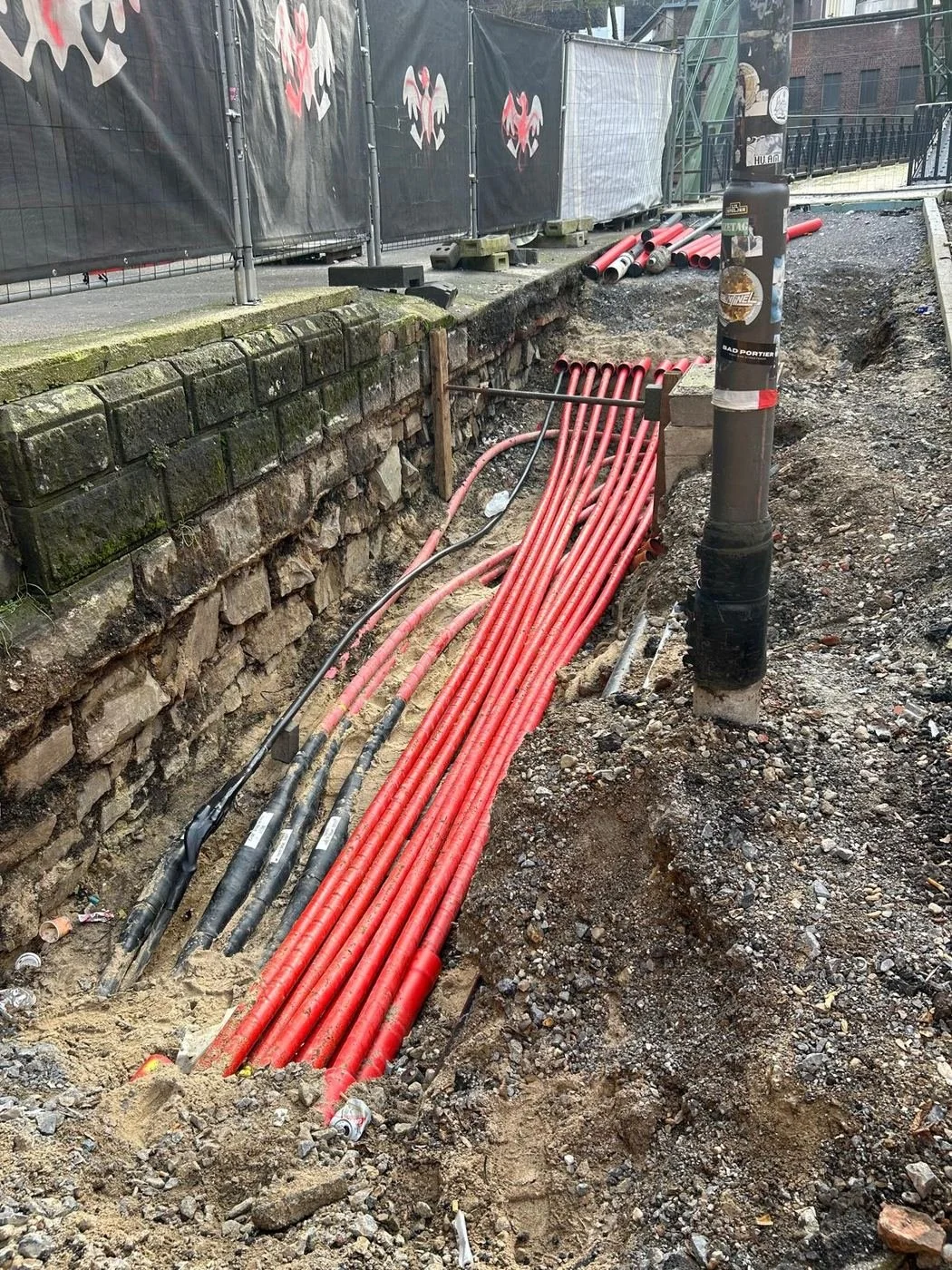 Offener Kabelgraben mit vielen roten Kabelschutzrohren an einer Natursteinmauer in Wuppertal