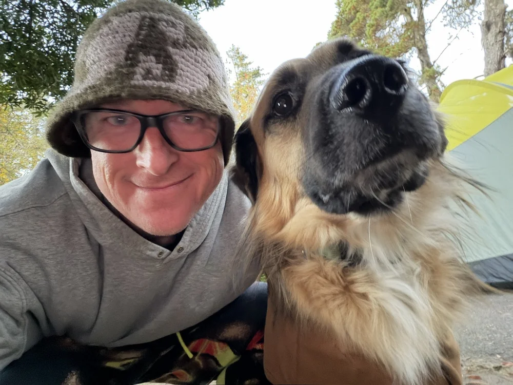 Mike grinning in a knit bucket hat as Atlas pushes his nose into the camera, yellow tent behind