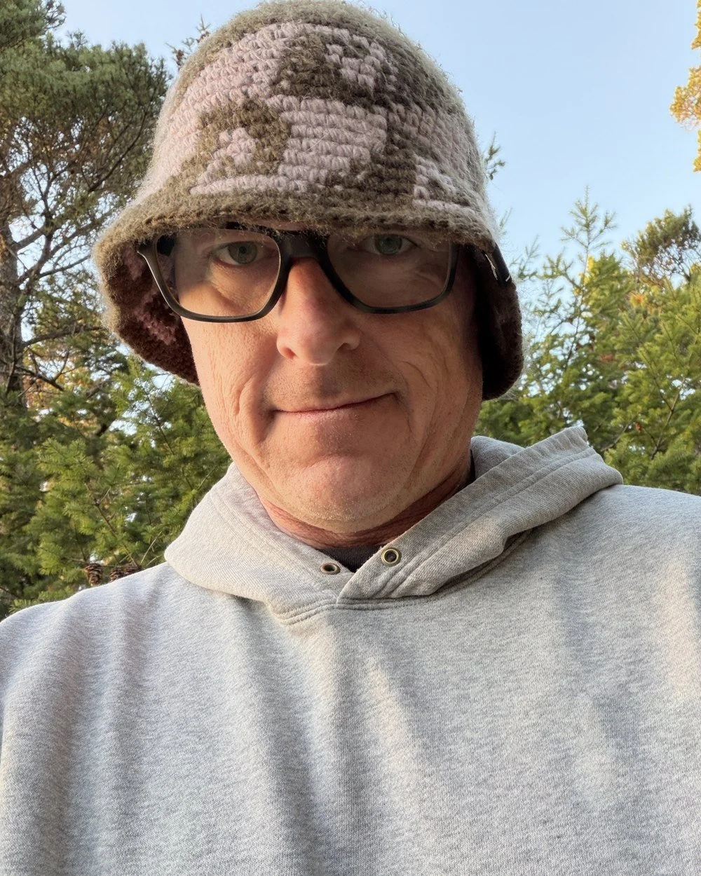 Mike in a knit bucket hat and grey hoodie at a coastal campsite, fall trees and blue sky behind him