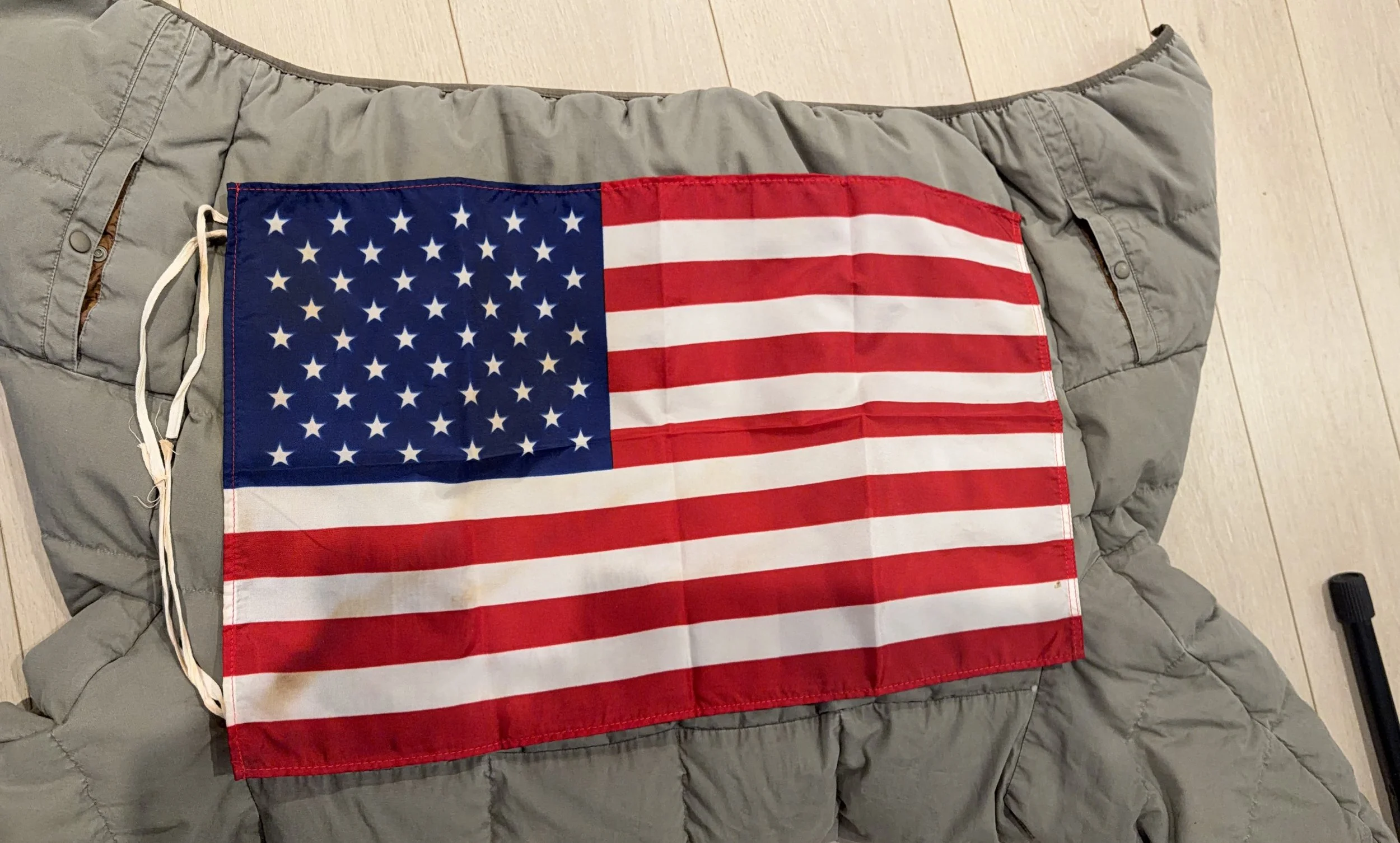 A small American flag with white ties, laid on a gray down jacket, with faint orange stains on the white stripes