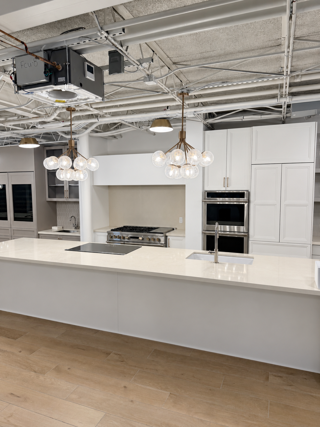 White kitchen cabinetry, double wall ovens and a long island beneath globe pendants in the TLTH showroom.