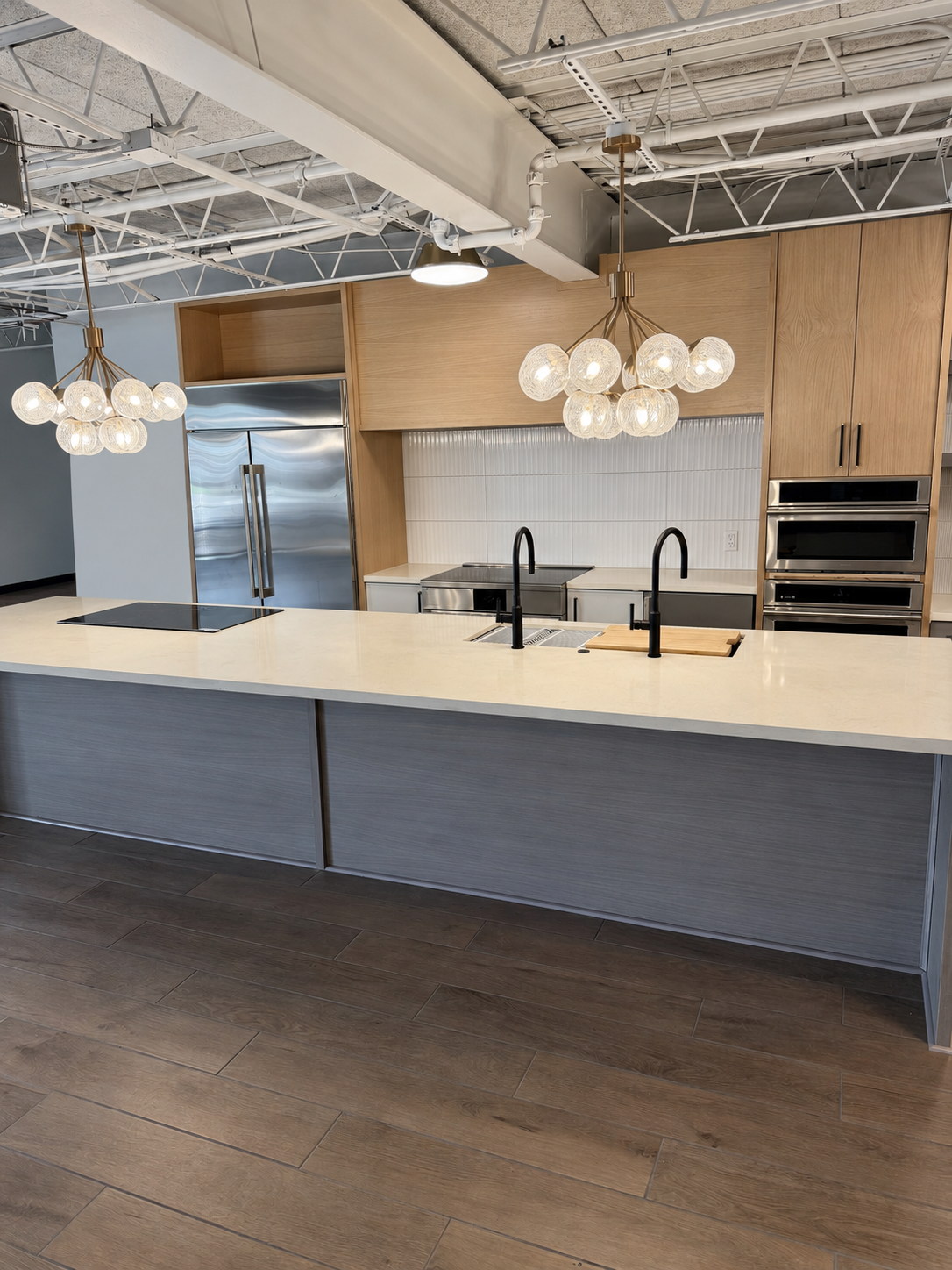 Wide view of oak kitchen cabinetry, stainless refrigeration and a gray island with dual faucets at TLTH.