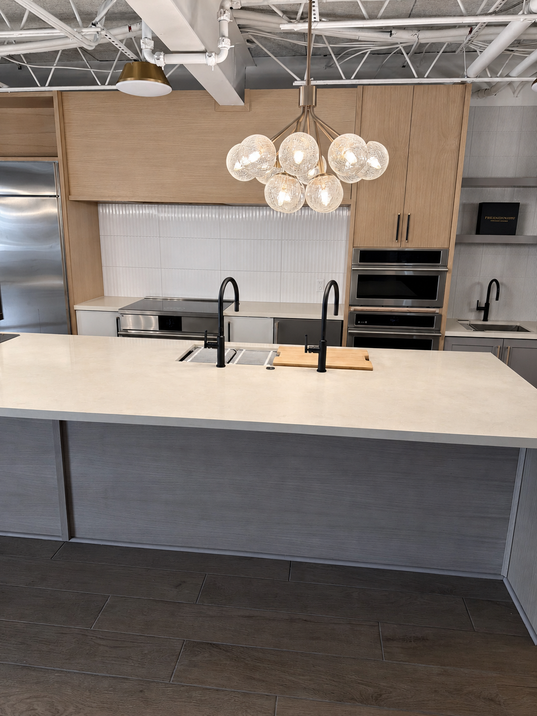 Oak wall cabinetry, a gray workstation island and dual black faucets at The Luxury Tree House showroom.