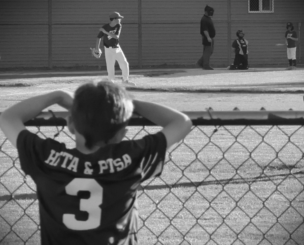 Black-and-white photograph of a young boy viewed from behind as he leans against a chain-link fence and watches a baseball game.