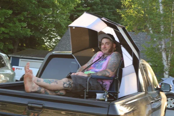 A tattooed man relaxing barefoot in a folding chair in the bed of a pickup truck beneath a large umbrella.